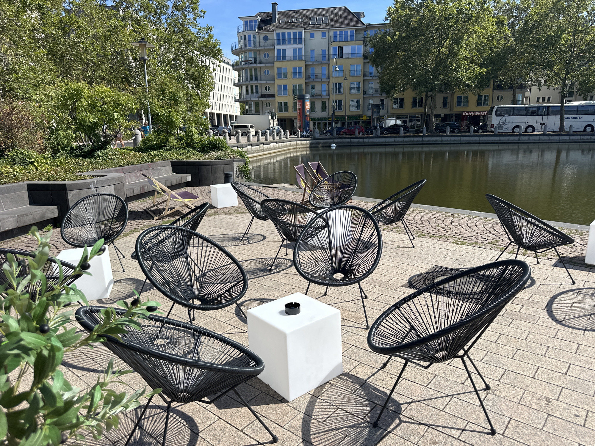 The image depicts an outdoor seating area adjacent to a body of water, likely a canal or pond, in the city of Cologne, Germany. The foreground features several black, woven patio chairs arranged on a paved surface. A white, cube-shaped side table with an ashtray on top sits in the center of the chairs. To the left, a potted plant with green leaves and black olives is visible. In the middle ground, a few lounge chairs are placed near the water's edge, and a low stone wall with built-in seating is present. The background reveals modern buildings, some with balconies and numerous windows, lining the opposite side of the water. Trees with full green foliage are scattered throughout the scene. A white bus with the text "VAN KOOTEN" is parked along the waterfront, suggesting a commercial or transport hub. The sky is clear and blue, indicating fair weather. A few people are visible in the distance, walking near the buildings and trees, but no specific interactions are apparent. The overall impression is a public outdoor space designed for relaxation and enjoying the urban waterfront.