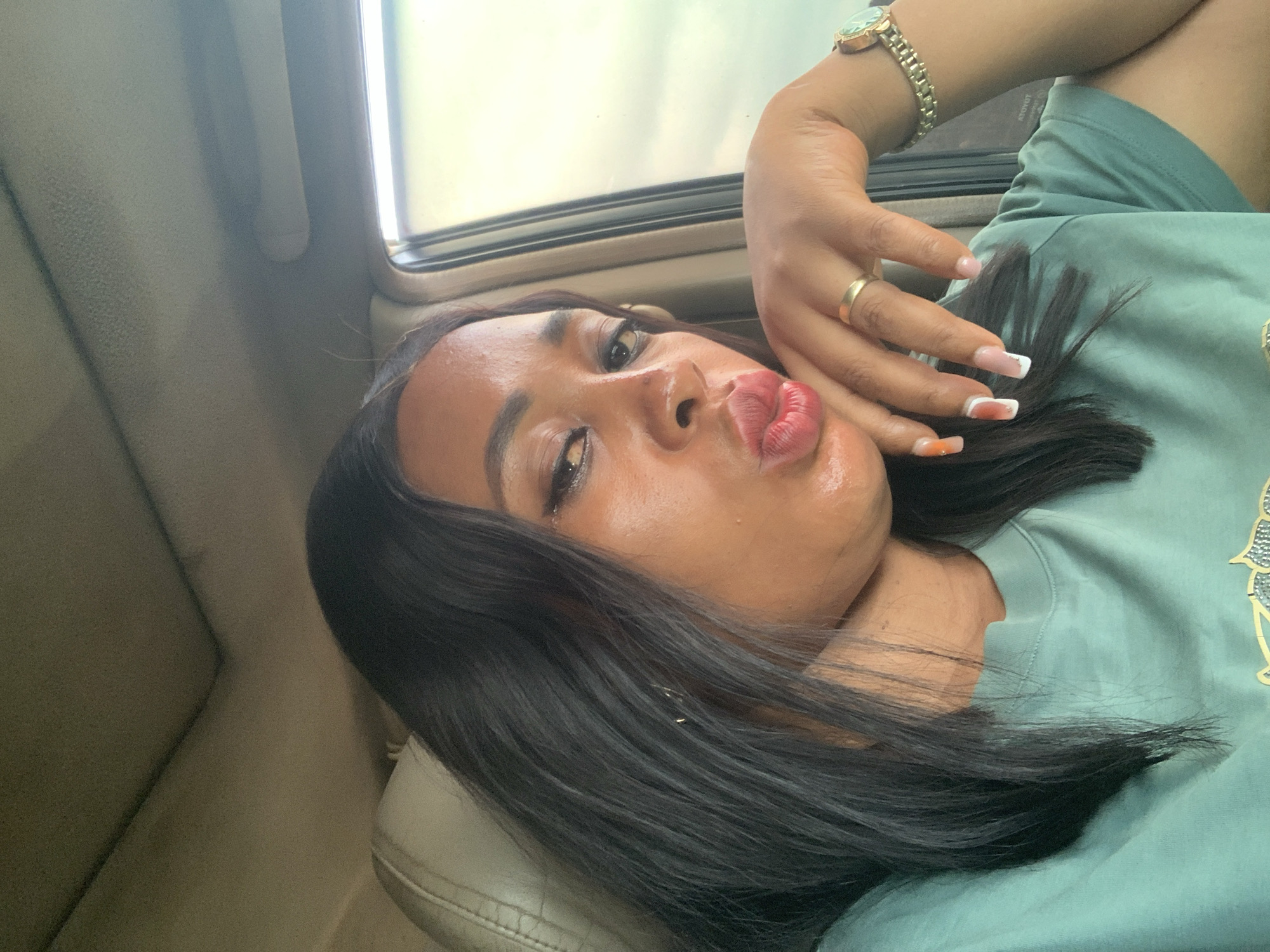 A woman is reclining in what appears to be a car. Her black hair is styled and swept to the side. She wears a green t-shirt, a gold watch, and a gold ring. Her nails are manicured with a white tip design. She is puckering her lips for a photo. The interior of the vehicle is a light tan color. There is a window visible in the background. The location of Sabon Tasha, Nigeria is not apparent in the image.