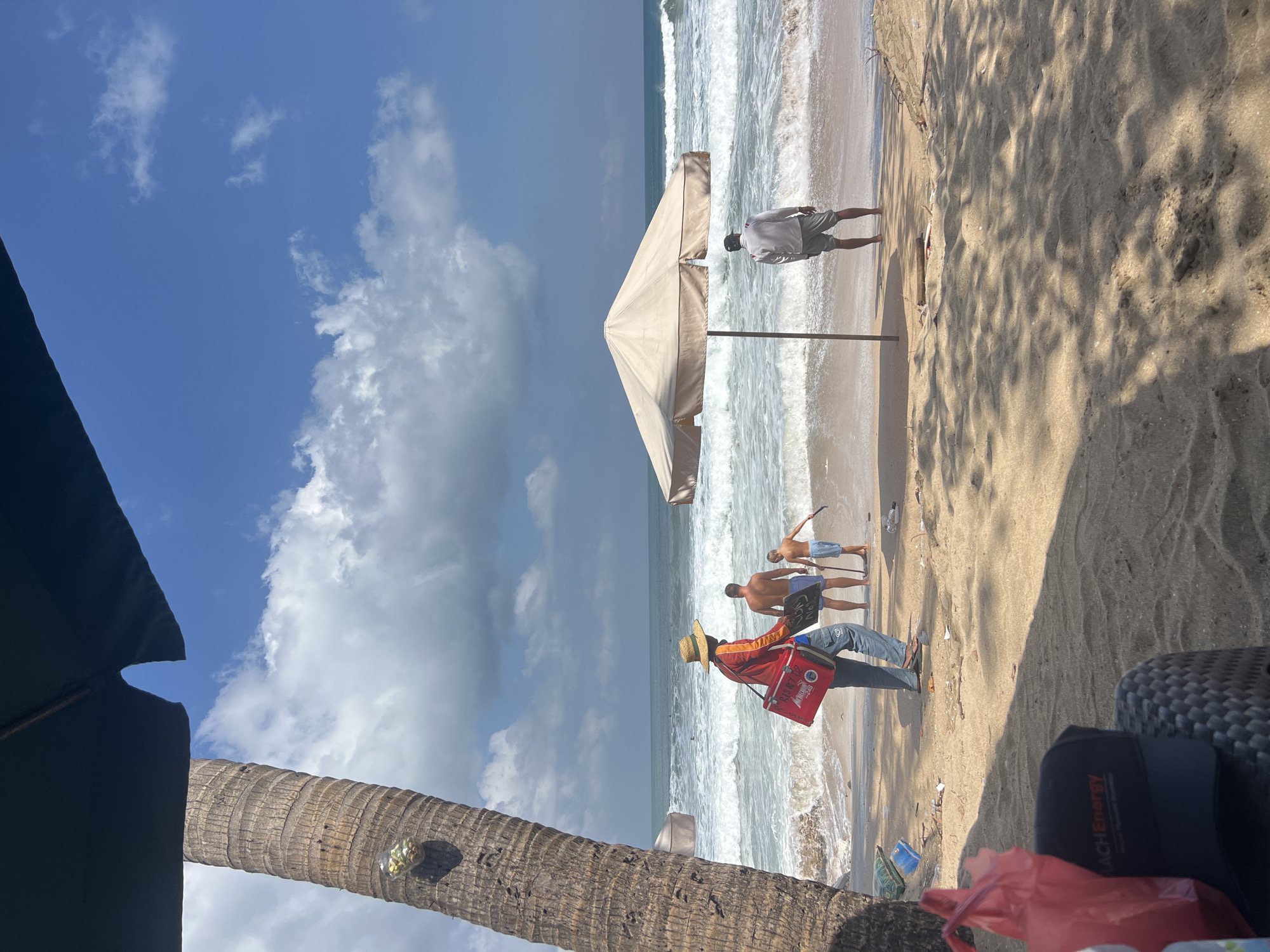 On a sandy beach in Kuta, Indonesia, under a bright blue sky with scattered clouds, two adult males and a young boy stand near the ocean. The sea is rough, with white-capped waves rolling onto the shore. To the left, a large palm tree stands tall. In the foreground, a tan beach umbrella provides shade. A person in a straw hat and blue jeans walks towards the water, carrying a red cooler bag and holding two signs. Another adult male, shirtless and wearing grey shorts, stands facing the ocean under the umbrella. The young boy, wearing blue shorts, is near the water's edge, holding a stick. The beach is littered with some trash, including a red plastic bag and a blue plastic container.