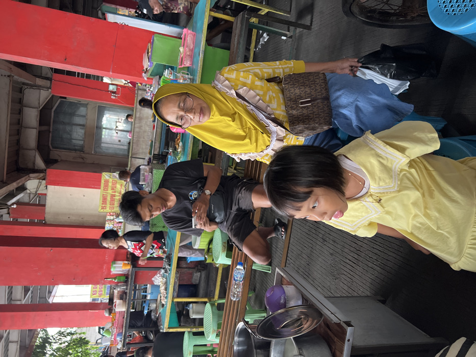 The image depicts a bustling food stall area within a market in Nganguk, Indonesia. In the foreground, a young girl, approximately 4-6 years old, with short black hair and wearing a yellow dress with a white collar, is seated on a blue plastic stool, looking towards the right with her mouth slightly open. Beside her, an older woman wears a yellow hijab, glasses, a yellow patterned long-sleeve top with ruffled trim, and a blue skirt. She carries a brown shoulder bag with a repeating pattern and holds a black plastic bag in her left hand, looking forward.

Behind these two individuals, a young man with dark hair, a black t-shirt with a white logo, dark shorts, and black flip-flops is seated on a wooden bench. He is focused on a white smartphone held in both hands. A clear plastic water bottle rests on the bench beside him. In the midground, several food stalls are visible, characterized by light blue and yellow counters displaying various items, some wrapped in aluminum foil, along with plastic containers and bottles. One yellow sign with red lettering advertises "ES DAWEF / CENDOL" and "ES GEMPOL & PLERET." A woman in a patterned top stands behind one of these stalls.

The structural environment features large, square red pillars supporting a high ceiling with exposed beams. The flooring beneath the seated man is concrete with visible grooves. Several green plastic stools are scattered around. Additional indistinct figures are present in the background, seated or standing, contributing to the busy atmosphere of a public eating area during daylight hours.