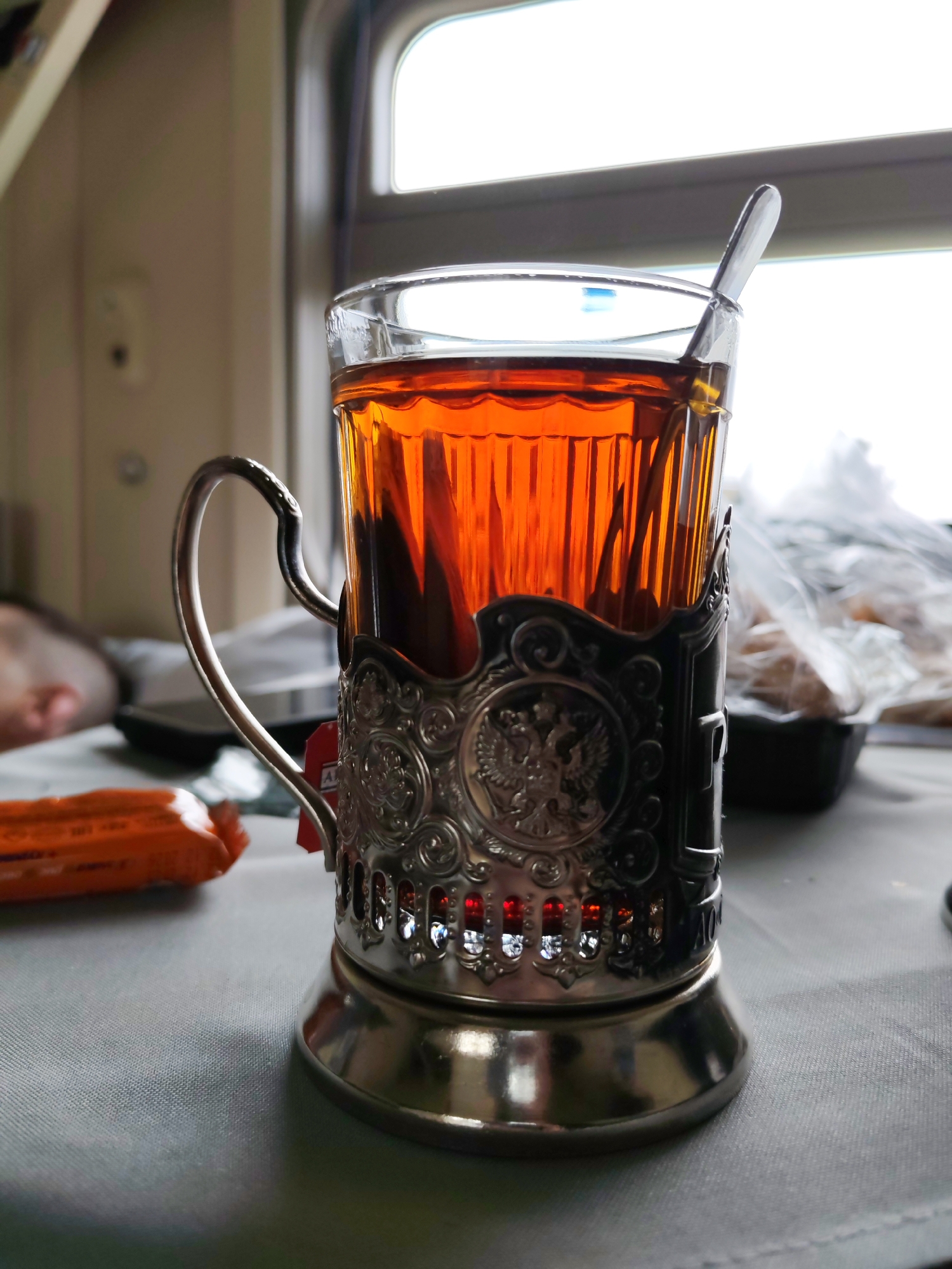 The image presents a close-up of a glass of amber-colored tea held within a traditional, ornate silver-colored podstakannik (metal tea glass holder). A metal spoon is placed inside the tea. The podstakannik is intricately designed with decorative patterns and prominently features the double-headed eagle, the coat of arms of Russia, as a central motif. A red tag, likely from a tea bag, hangs from the holder, with the words "CEYLON TEA" visible, and possibly part of "Ahmad Tea" above it.
The scene is set inside what appears to be a train car, suggested by the shape of the window visible in the background and the common use of podstakanniki on Russian railways. Bright daylight is evident through the window, indicating it is daytime. To the left in the soft-focus background, a person's head is faintly visible, resting on a pillow or seat, suggesting they are asleep or relaxing during the journey. The tea glass and holder rest on a light-colored, possibly grey, patterned surface, likely a tablecloth. Other blurry items in the background include an orange-wrapped snack or candy bar and a black container with food items wrapped in clear plastic, typical for travel provisions. The image conveys a serene moment of travel and refreshment within Russia, specifically around Borduki.