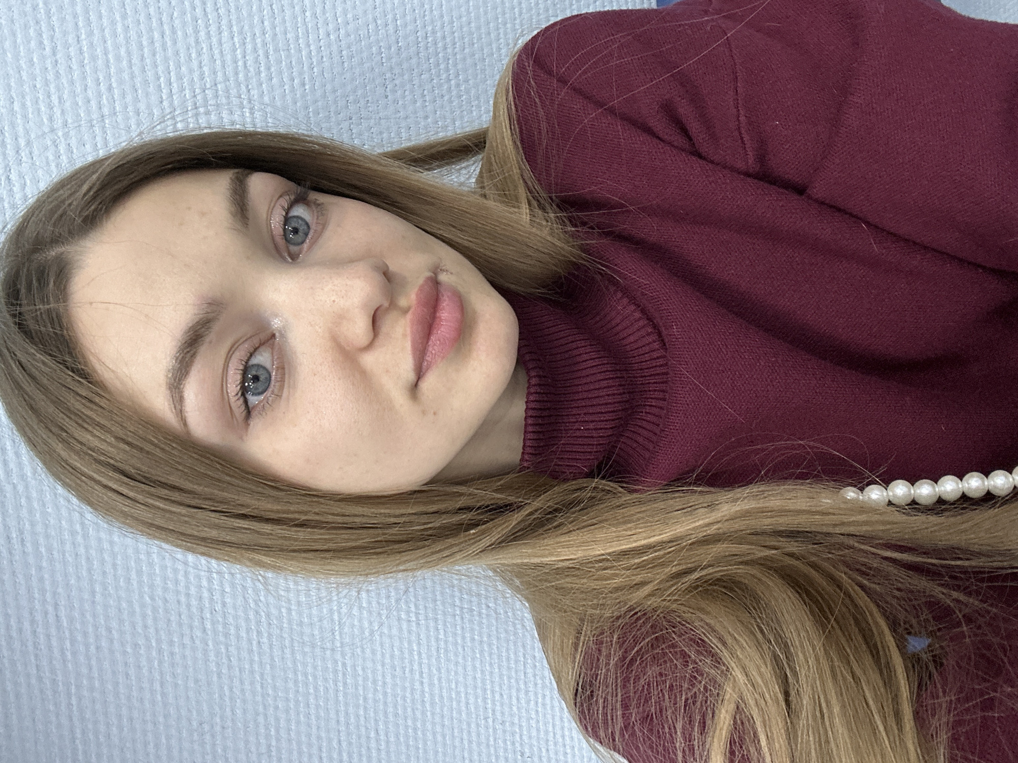 A woman with fair skin and light brown hair is pictured. She has blue eyes and pink lips. She is wearing a burgundy turtleneck sweater and a pearl necklace. The background is a light blue textured surface. The image is taken at an angle and is slightly rotated. There is no specific evidence to link the location to Kazan, Russia.