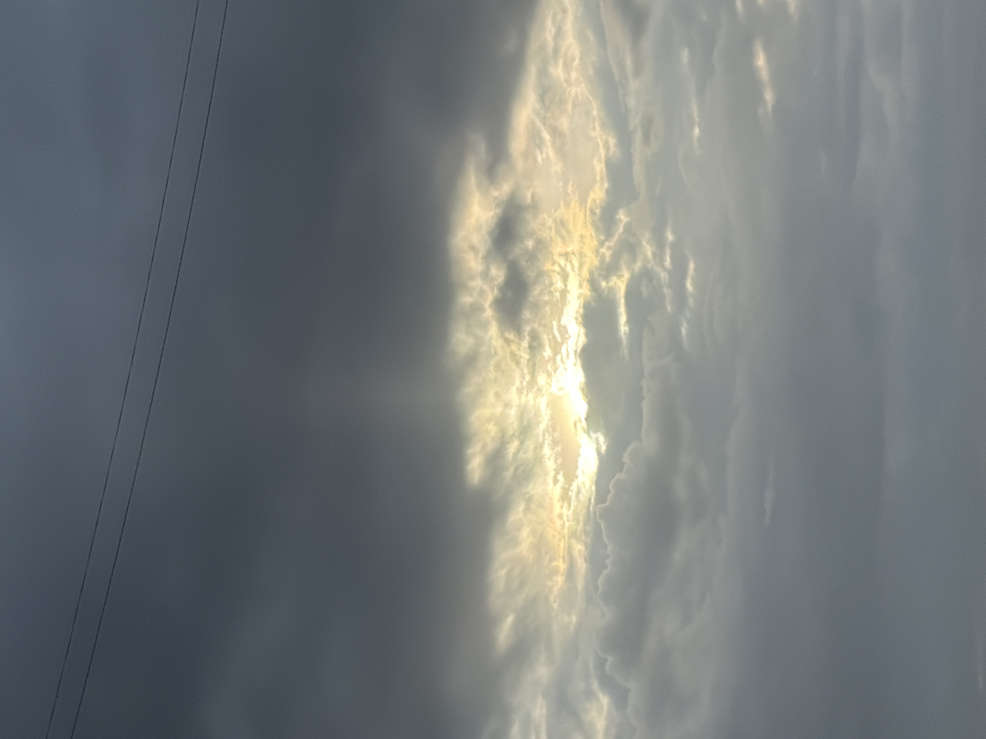 The image shows a cloudy sky with a patch of bright sunlight breaking through the clouds. Two electrical wires are visible in the upper left of the frame. The image has been rotated counterclockwise. The specific location in Chaikovsky, Russia cannot be determined from the image alone. No people, subjects, objects, actions, or interactions are explicitly shown in the image beyond the elements mentioned. The environment is predominantly atmospheric, characterized by clouds and light.