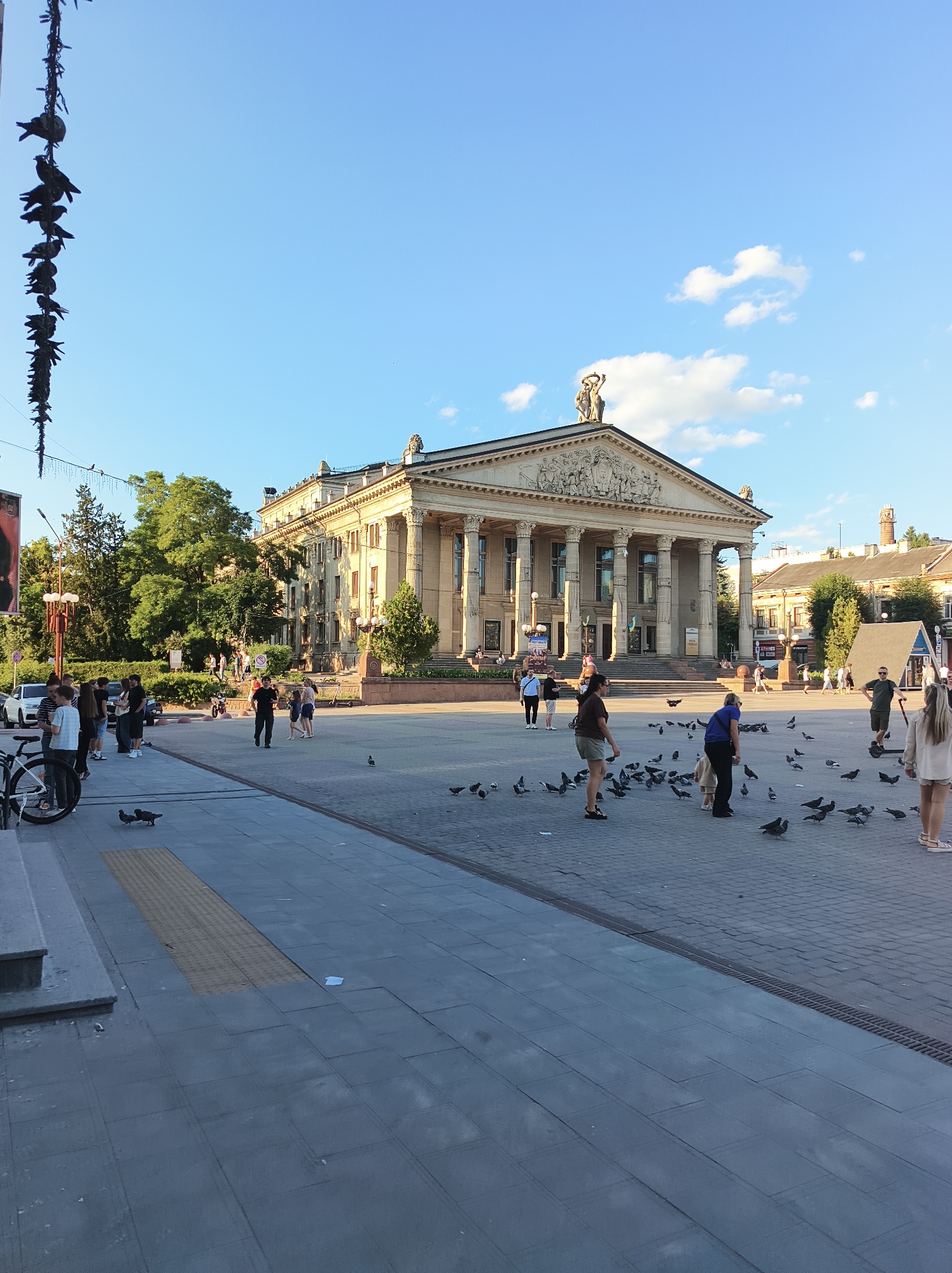 The image depicts a public square in front of a large neoclassical building, likely a theater or government building, in the city of Ternopil, Ukraine. The building features a grand portico with columns and intricate sculptures on its pediment. The square is paved with gray stone tiles and is populated by a number of people and a significant flock of pigeons.
In the foreground, several people are standing and walking on the paved square. Some are interacting with the pigeons, while others are engaged in conversation. A bicycle is parked on the left side of the image near a set of stairs leading up to a building. The ground in the foreground shows some wet patches, suggesting recent rain or cleaning.
In the middle ground, more people are scattered across the square, some are feeding or observing the pigeons. A small, A-frame structure is visible to the right of the main building. A number of cars are parked on a street adjacent to the square.
The background features lush green trees, other buildings, and a clear blue sky with scattered white clouds. The overall scene conveys a sense of everyday urban life, with people enjoying the public space amidst the avian presence. The building's architecture suggests a historical and culturally significant location within Ternopil.