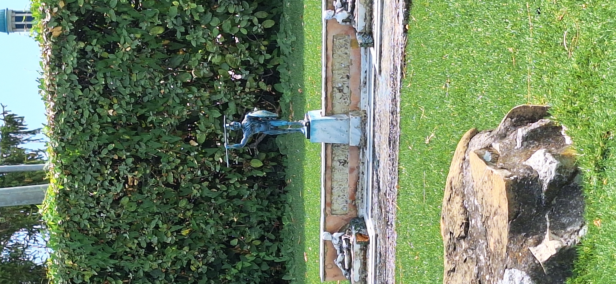 The image captures a bronze statue of a warrior holding a spear and shield, positioned centrally on a light blue pedestal. The statue is located outdoors in a garden setting, with a dense green hedge forming the backdrop. In front of the hedge, a low, decorative brick wall is visible, featuring a frieze with intricate carvings. To the left of the main statue, on a lower level, a reclining stone statue is partially visible. The foreground is dominated by a large, rough-textured rock on the left and a well-maintained green lawn covering the rest of the visible ground. The overall scene suggests a historical or artistic garden.