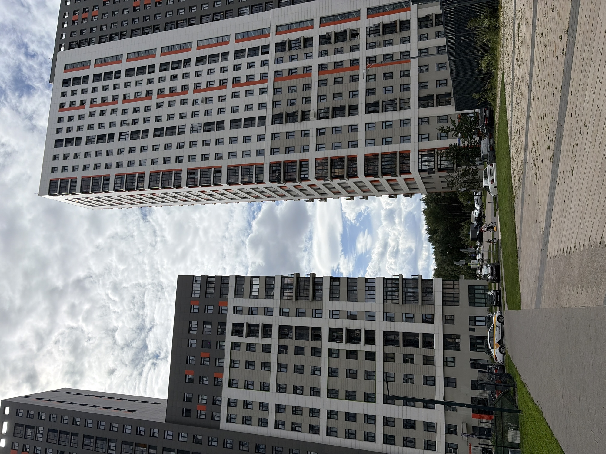The image shows a view of two modern high-rise residential buildings in Kotelniki, Russia, against a cloudy sky. The buildings are tall and rectangular, with many windows and balconies. The building on the left is primarily gray and white with orange accents, while the building on the right is white with orange vertical stripes. In the foreground, there is a paved pathway and a grassy area. Several cars are parked in front of the buildings, including a white taxi with yellow markings. A few people are visible in the distance, including individuals riding bicycles. The scene appears to be a residential complex in a suburban or urban area.