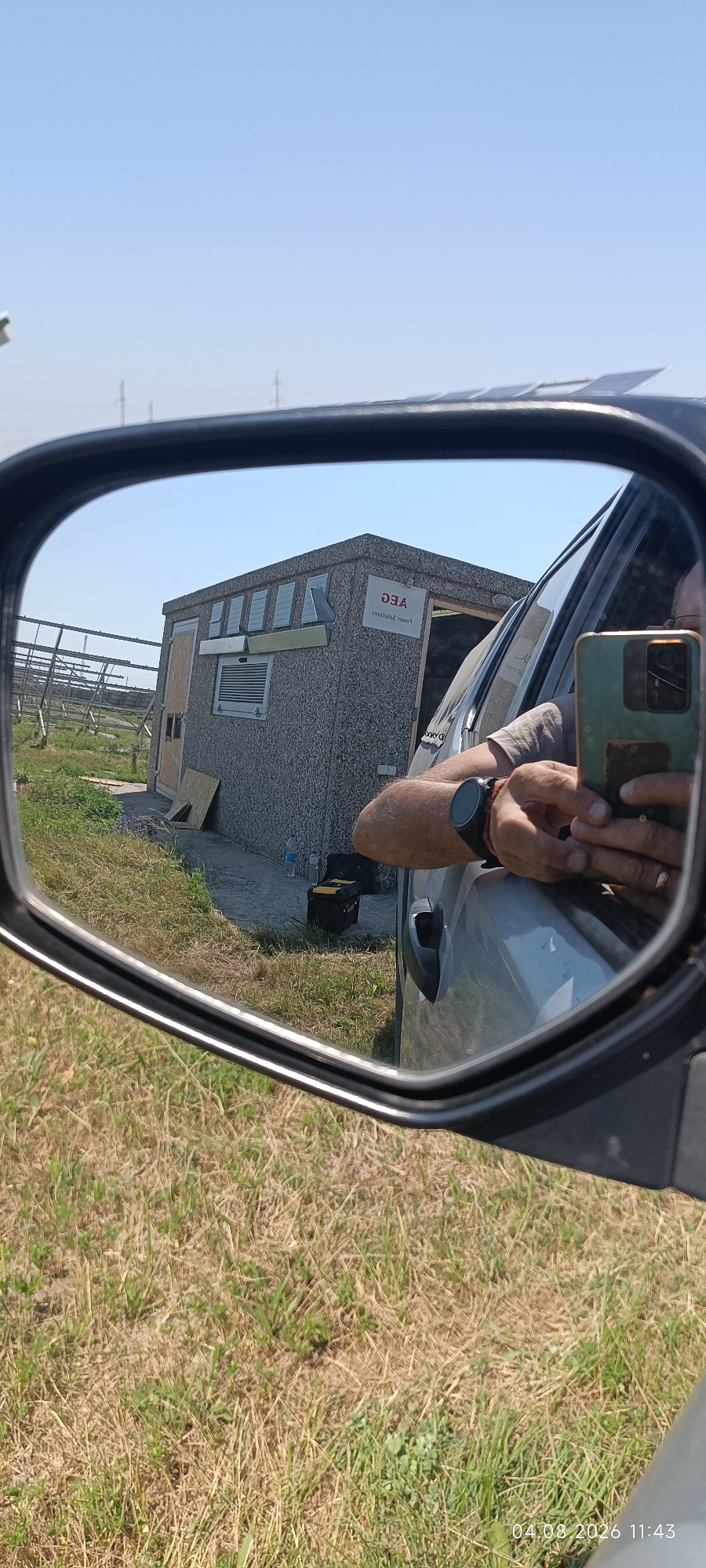 The image, taken in Starokozache, Ukraine, reflects a scene in a side-view mirror of a vehicle. The mirror shows a small, utilitarian building with a sign that reads "AEG Power Solutions" in reverse. The building appears to be constructed from textured, gray material and has several windows or vents. In front of the building, there are some wooden boards and a yellow tool case. Beyond the building, a field with solar panel arrays is visible. The reflection also shows the arm and hand of a person holding a smartphone, presumably taking the photo. The environment outside the vehicle appears to be a dry, grassy field under a clear blue sky. The timestamp on the image indicates it was taken on August 4, 2026, at 11:43 AM.