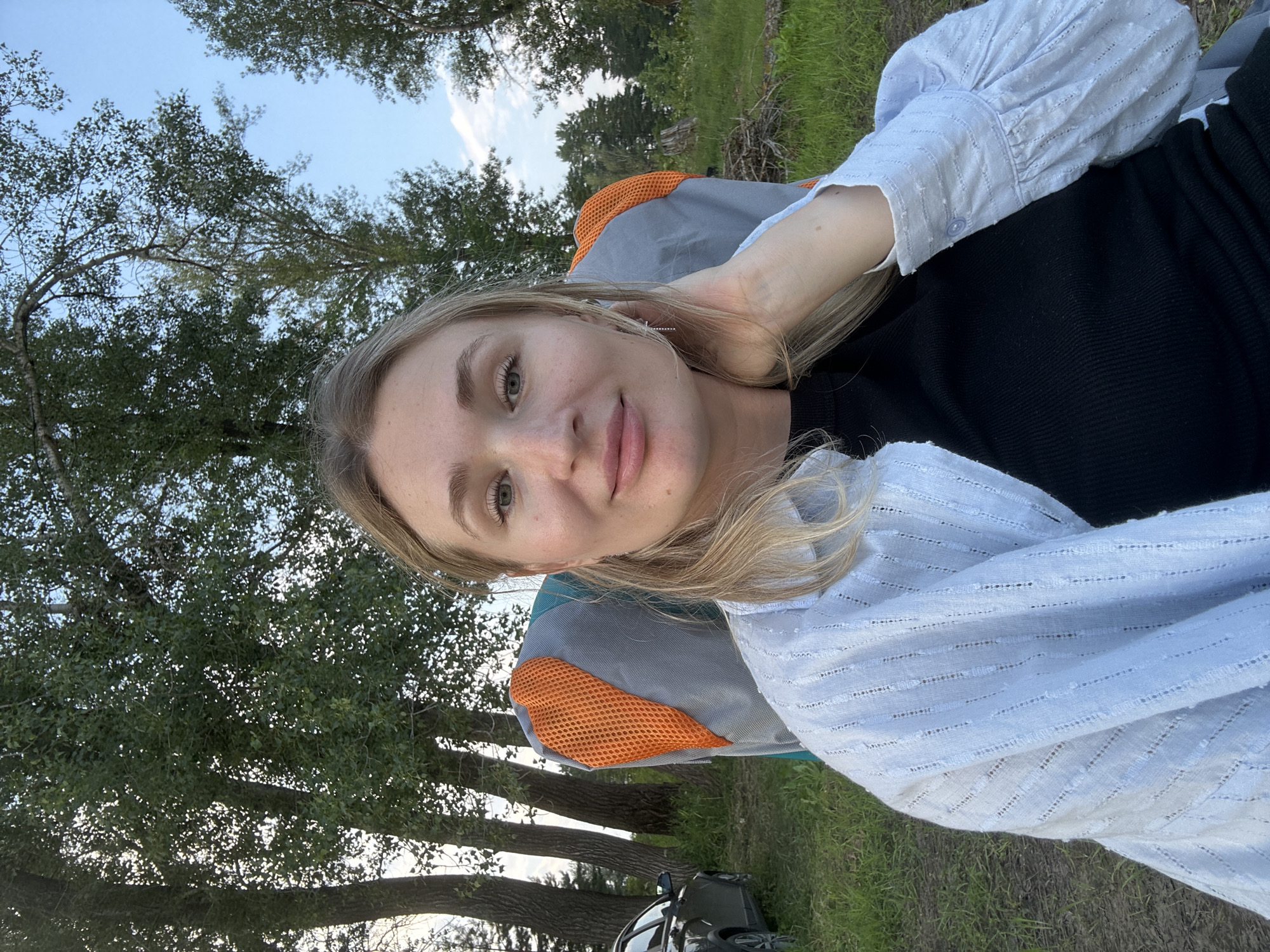 A young woman with blonde hair is seated in a camping chair, with her right hand touching her head. She is wearing a light blue, open-weave shirt over a black top. The background shows a natural, outdoor setting with trees and grass, suggesting a park or campsite. To the left, a dark-colored car is parked on a dirt path. The image appears to have been taken in or around Noviy, Russia, given the context provided.