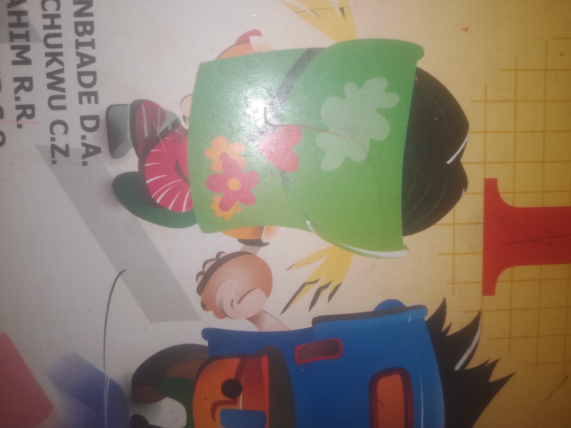The image shows a portion of a colorful children's book or educational material, featuring cartoon characters. On the left side, partially visible text reads "NBIADE D.A. CHUKWU C.Z. AHIM R.R.". Prominently displayed is a character's head covered by a green sheet adorned with light green leaf shapes and pink and orange flowers. Below this, a character's hand is visible, reaching out. At the bottom, a blue toy train with orange and black detailing is depicted. The background is a plain white surface with a yellow grid pattern to the right, suggesting a table or floor. There are no discernible elements in the image that would allow for specific location identification in Jalingo, Nigeria.