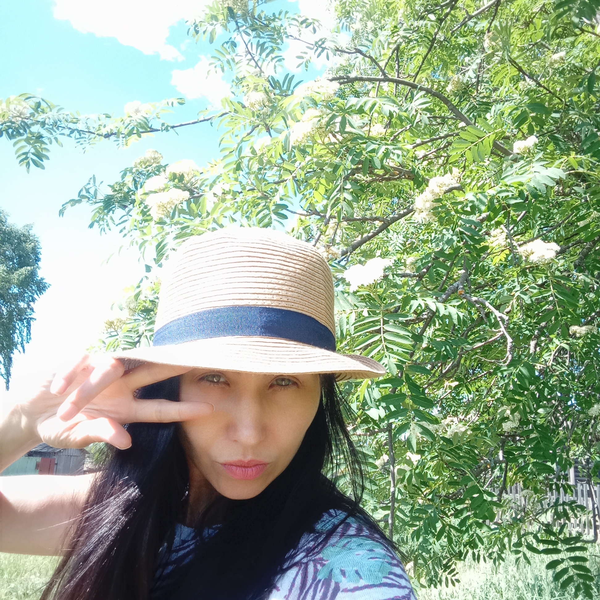 A woman in Буренка, Russia, poses for a selfie in a garden or yard. She wears a light brown straw hat with a dark blue band and has long dark hair. She is wearing a top with a blue and purple leaf-like pattern. She holds up her hand in a "peace" sign near her face. Behind her is a tree with green leaves and small white flowers. The sky is blue with some scattered clouds. A glimpse of a wooden fence and a small building is visible in the background. The scene is filled with natural light.