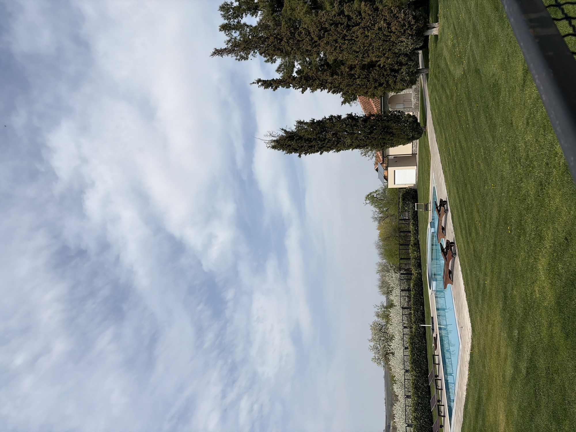 The image shows a landscape view in Topola Municipality, Serbia. In the foreground, a swimming pool with lounge chairs beside it is surrounded by a well-maintained lawn. A building is partially visible beyond the pool and lawn. Trees line the top of the image, and the sky is filled with clouds.
