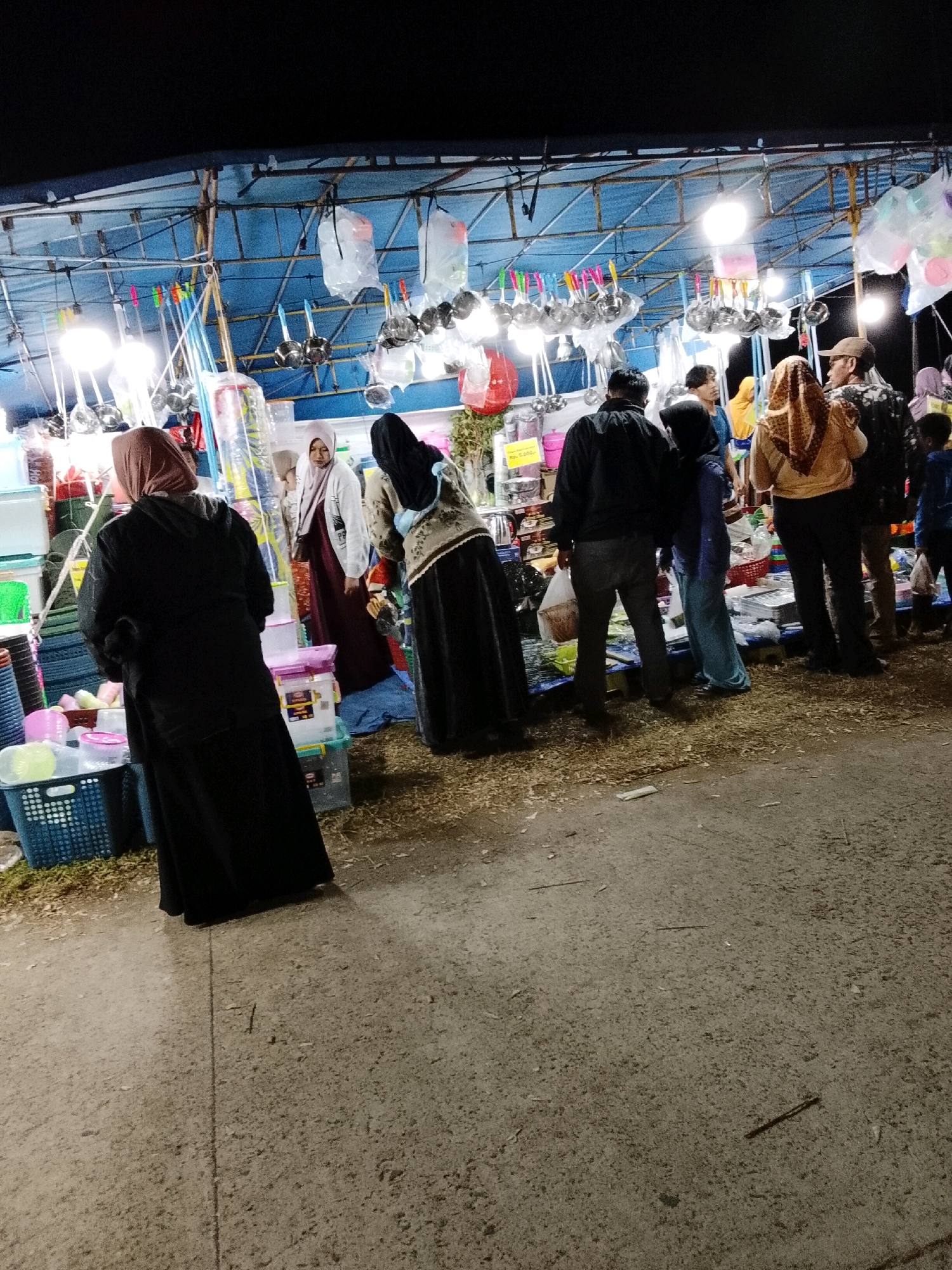 The image depicts an outdoor market scene at night in Padang Dalom, Indonesia. A stall is set up with various items for sale, including kitchen utensils like ladles and sieves hanging from the ceiling, as well as plastic containers and baskets. Several people are browsing or shopping at the stall. The ground appears to be a mix of dirt and loose gravel. Lighting comes from overhead bulbs, creating a somewhat dim and atmospheric environment. The individuals present are dressed in casual clothing, with some wearing headscarves. The overall impression is of a bustling local market at night.