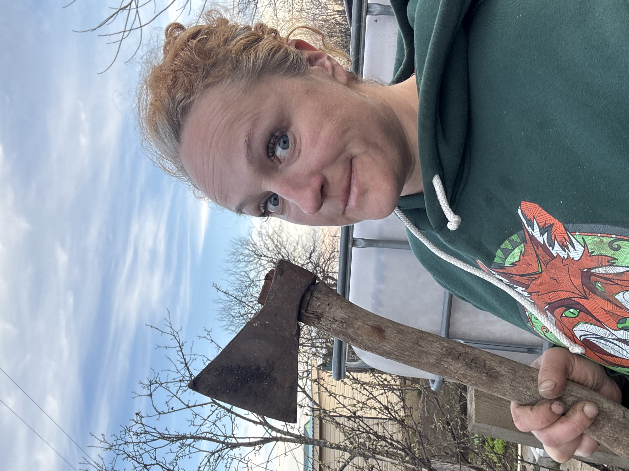 A woman with fair skin, blue eyes, and light reddish-brown curly hair pulled back is depicted in a vertical, slightly low-angle shot. She looks directly at the camera with a subtle smile. She wears a dark green hooded sweatshirt featuring a vibrant, stylized graphic of a fox-like creature with green eyes on the front, and a white drawstring at the neck. Her left hand, showing some dirt under the fingernails, firmly grasps the wooden handle of an old, rusted axe. The axe head, made of dark metal, shows signs of significant wear and rust, and the wooden handle appears aged.
The background reveals a clear, light blue sky with delicate, wispy clouds, indicating daytime. Bare tree branches, suggesting late winter or early spring, are visible across the upper and left portions of the frame. On the right, part of a large white plastic container, possibly an IBC tote, is visible. Indistinct building structures can be faintly seen in the far background. The scene takes place outdoors, in what appears to be a semi-rural setting in Chaikovsky, Russia. There is no visible text in the image.