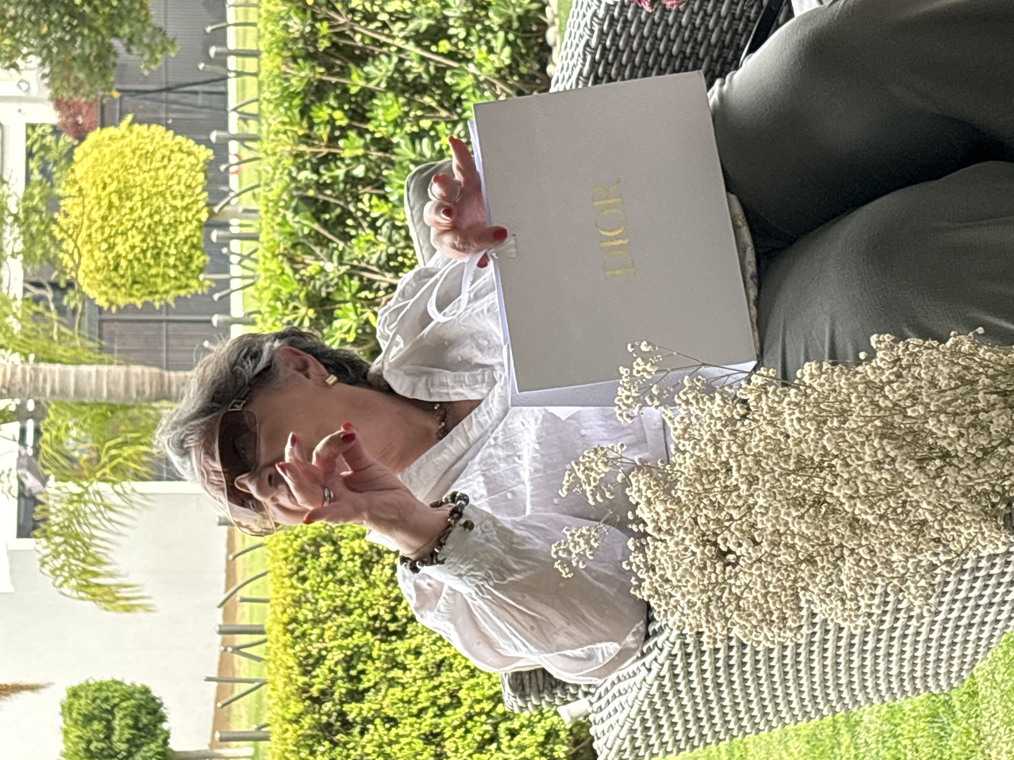 The image shows an older woman sitting in what appears to be a garden or outdoor patio setting. She is wearing sunglasses and a white blouse, and holding a white box with "DIOR" printed on it in gold lettering. There is a bouquet of small white flowers next to her on a woven chair. The background includes green foliage, including a manicured hedge and a round shrub. Although there is no way to be 100% sure without additional information, the location is reported to be San Bernardino Tlaxcalancingo, Mexico.