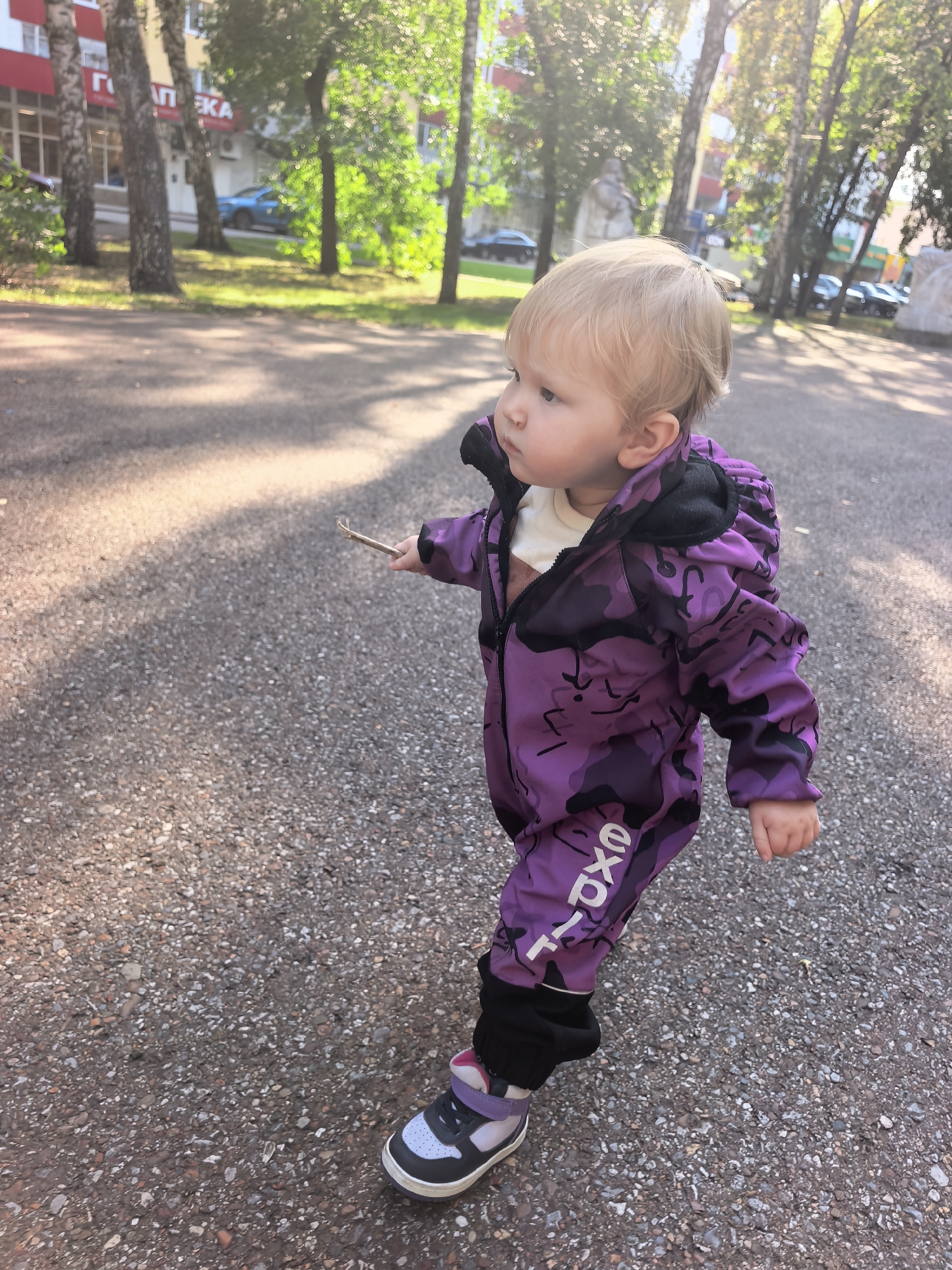 A toddler with blonde hair is walking on a paved path outdoors. The child is wearing a purple camouflage hooded jumpsuit with black trim and white lettering on the leg, and purple and white sneakers. The child is holding a small twig in their right hand. In the background, there are trees, a building with a red and white facade, and parked cars. The setting appears to be a residential area in Sterlitamak, Russia, as indicated by the Cyrillic lettering on the building. The sunlight is dappled, suggesting it is daytime.