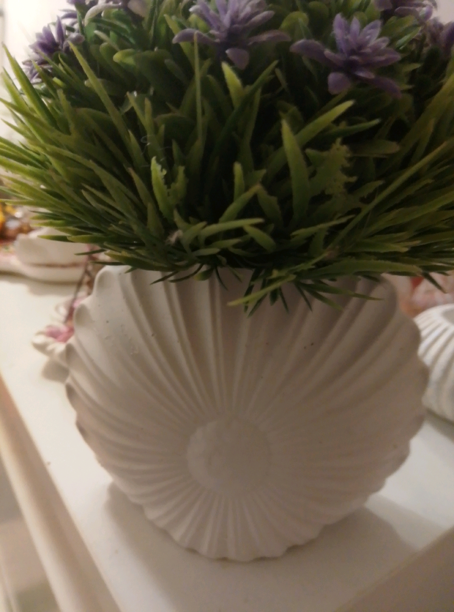 The image shows a close-up of a white, seashell-shaped planter filled with artificial green grass and purple flowers. The planter has a ribbed texture radiating from the center. The background is blurred and appears to be an indoor setting with a white surface that could be a shelf or table. There are no people visible in the image. The location of this scene in Wedel, Germany, is not discernible from the visual information provided.