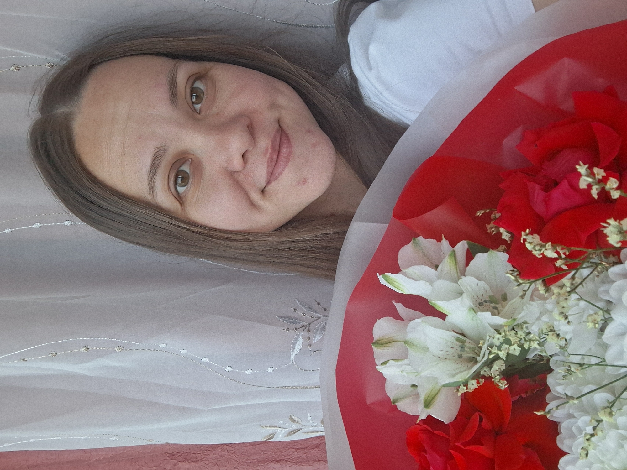 A woman with long brown hair and light skin is holding a bouquet of red and white flowers. The flowers are wrapped in red paper. The woman is wearing a white shirt. The background is a white curtain with a floral pattern. The location is likely in Chaikovsky, Russia, based on the user's prompt.