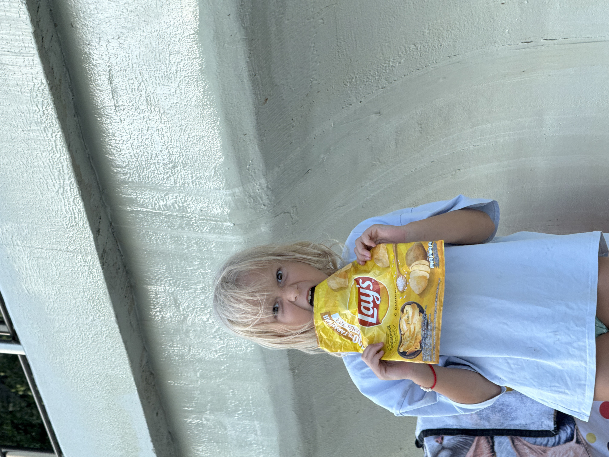 A young child with blonde hair stands outdoors, biting into a yellow bag of Lay's potato chips. The child is wearing a light blue t-shirt and has a mischievous expression. Behind the child is a textured, pale green wall, possibly a concrete structure. A railing is visible at the top of the image. The lighting suggests it is daytime. The specific location in the city of Unknown, Ukraine cannot be determined from the visual information provided.