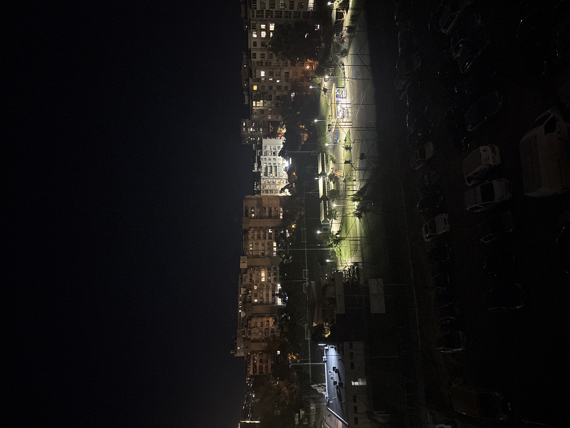 This is a nighttime, aerial, vertical photograph of a city scene. The image shows a row of apartment buildings on the left side, with many windows illuminated, suggesting that people are inside. Adjacent to the buildings, on the right, is an outdoor basketball court with lights illuminating the playing area. Beyond the court, a parking lot is visible, filled with numerous parked cars. The scene is dark, indicating it is nighttime. There are no people or visible activity taking place on the basketball court or in the parking lot. The location and weather are unknown. There is no visible text in the image.