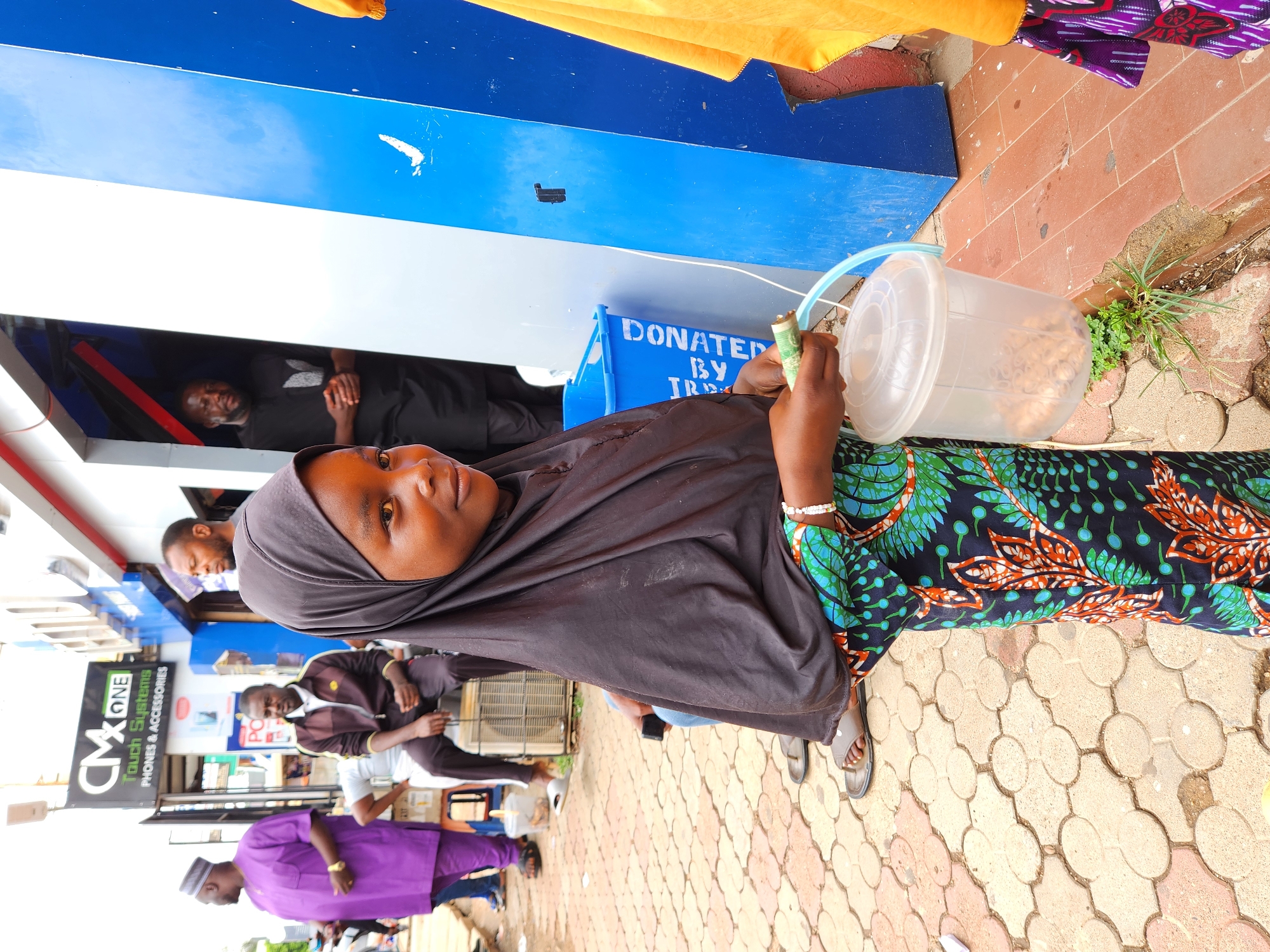 A street-level, medium shot shows a woman holding a clear plastic bucket with a blue handle in her right hand, which is folded to hold a rolled-up bill. The bucket contains a large number of what appear to be peanut balls. She is wearing a dark head covering and a green and black floral-patterned dress. Behind her are several people, including a man in a black shirt and a man in a purple shirt. A shop sign is partially visible in the background with the text "CM ONE TOUCH SYSTEMS PHONES & ACCESSORIES." The bucket she holds sits adjacent to the curb. The scene appears to be taking place in an urban environment, with a tiled walkway and various commercial buildings in the background. A donation box sits to the left of the subject. The background is visually cluttered.
