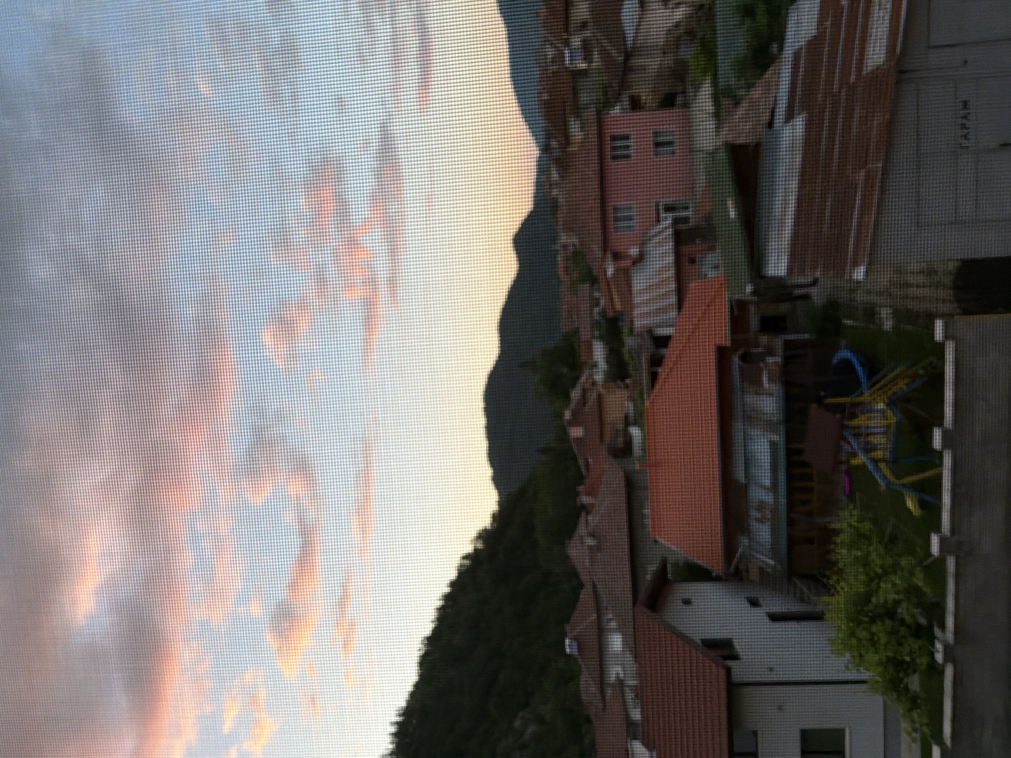 This image captures a residential area in Etropole, Bulgaria, at dusk. The foreground is dominated by the textured pattern of a screen, through which a cluster of houses with red tile roofs is visible. The houses vary in architectural style and color, with some painted in white and others in pink. A small playground with blue and yellow structures is situated in a yard. Beyond the houses, a dark, tree-covered hillside rises, leading to a mountain range in the background, silhouetted against a sky filled with soft, pastel-colored clouds. The overall impression is one of a quiet, suburban evening in a mountainous region.