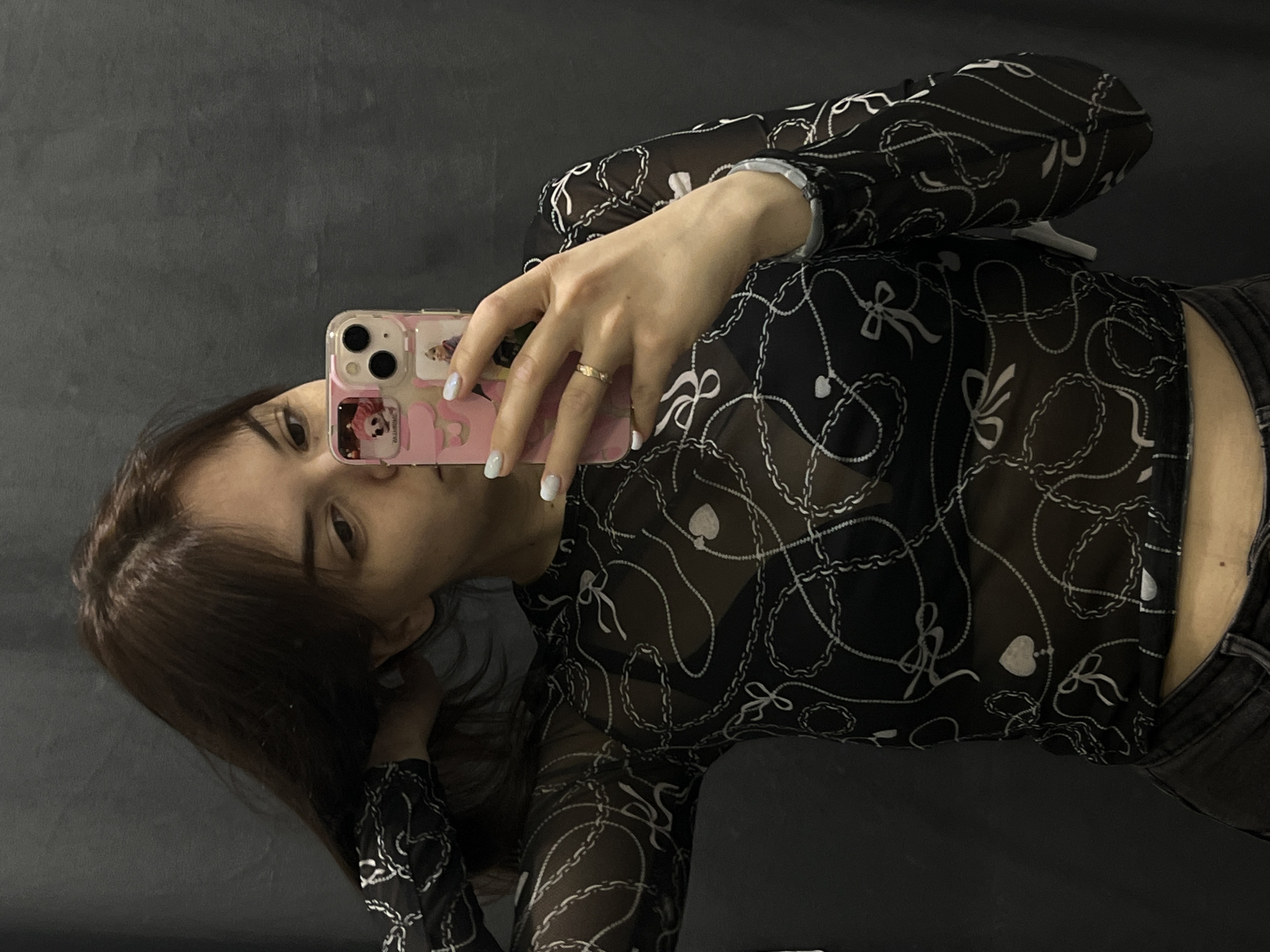 A young woman is taking a selfie in what appears to be an indoor setting. She is holding a pink smartphone with images on the case, wearing a sheer black top with a white chain and bow pattern, and a gold ring on her finger. The background is a plain gray surface. There is no indication of the image's location being Shushary, Russia.