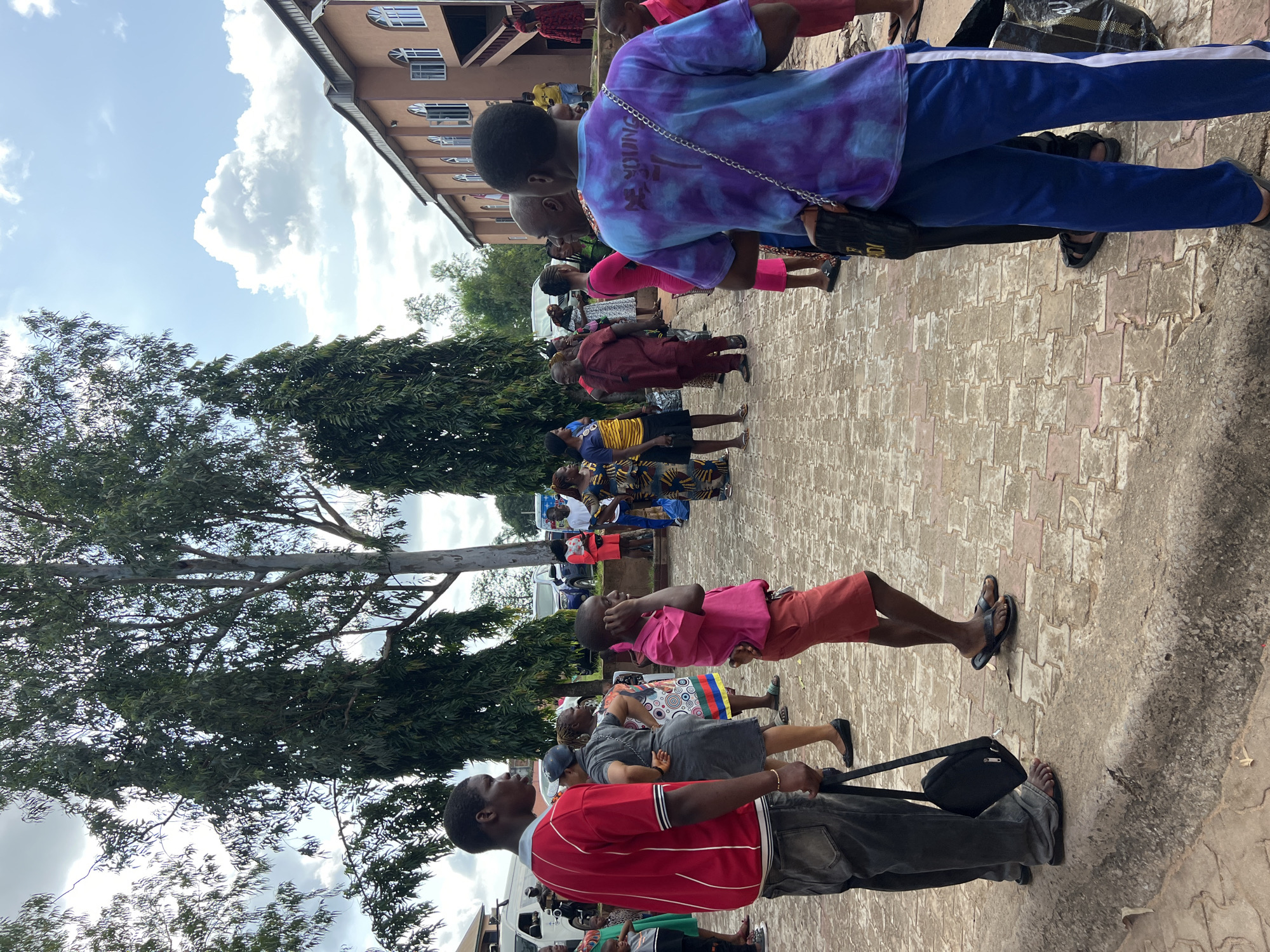 The image depicts an outdoor gathering of people in Nnewi, Nigeria, outside a building that appears to be a church or community hall, characterized by its reddish-brown facade and arched windows. Tall trees with lush green foliage dominate the background, with a cloudy sky above. The ground is paved with interlocking bricks.
In the foreground, a young boy in a pink shirt and red shorts stands with his hand to his ear. To his left, a man in a red shirt and grey jeans is seen from the side, holding a black bag. Several other individuals are scattered throughout the scene, some in brightly colored traditional Nigerian attire, while others wear more casual clothing. A group of people are conversing, and some appear to be observing an event or waiting. The overall atmosphere suggests a community event or a religious gathering.