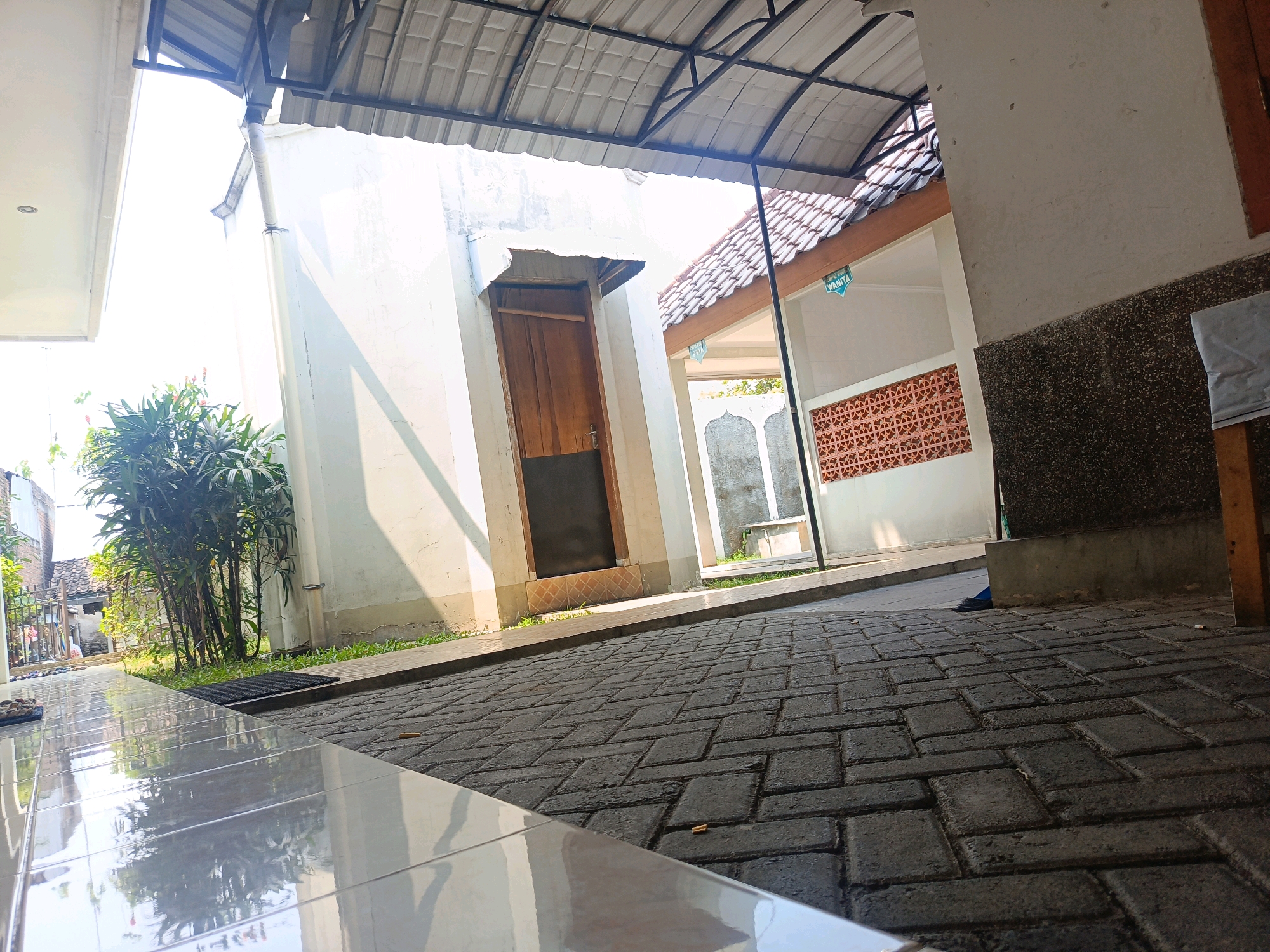 The image shows an outdoor courtyard area with tiled flooring in the foreground and brick paving further back. To the left, a white wall casts a shadow from a metal overhang above. A wooden door is visible on this wall. In the background, there is a building with a tiled roof, an open structure with patterned walls, and greenery. A blue object, possibly fabric, is on the ground near the building's foundation. The scene appears to be in a public or communal space, with a sign that reads "WANITA" indicating a women's facility. The overall lighting suggests it is daytime.