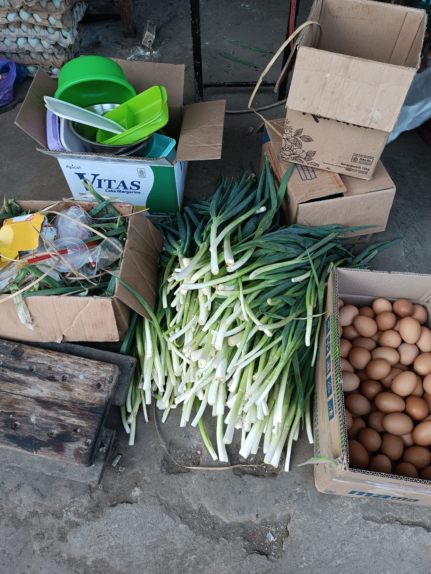 An outdoor market scene in Imogiri, Indonesia, displays an assortment of goods laid out on a concrete surface. In the upper left, a stack of pale blue egg cartons is visible. To their right, a large cardboard box overflows with various plastic and metal bowls and containers in shades of green, white, and purple. Adjacent to this, another cardboard box contains a large pile of green onions, their white bases and green stalks clearly defined. To the right of the green onions, a cardboard box is filled with numerous brown eggs. In the foreground, a weathered wooden plank rests on the concrete, and to its left, another open cardboard box holds a collection of discarded plastic cups, straws, and yellow packaging. The overall impression is one of a busy, perhaps slightly disorganized, market stall.
