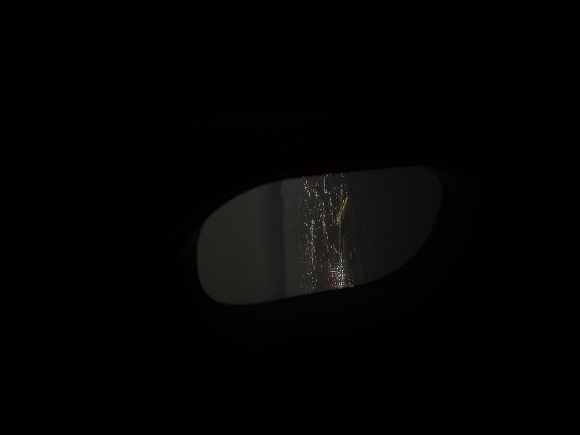 The image displays a nighttime urban scene viewed through a vehicle's side mirror. The majority of the frame is deep black, suggesting the dark interior of the car or very low ambient light outside the mirror's view. Centered within this darkness is an elongated, roughly oval-shaped reflection, which is the side mirror.
Within the mirror, a blurred and streaky cityscape is visible, illuminated by numerous small, bright artificial lights. These lights, predominantly yellow, white, and some red, are distorted by what appears to be rain or condensation on the mirror's surface, creating vertical streaks and diffused glows. This detail strongly indicates wet weather conditions. The lights suggest the presence of buildings, streetlights, and possibly traffic in an active urban environment.
The perspective is from inside a vehicle, looking out at the city at night. The scene captures a moment of quiet observation during wet weather in an urban area, consistent with the context of Huangpu District, China. There are no discernible human figures, specific landmarks, or visible text in the image.