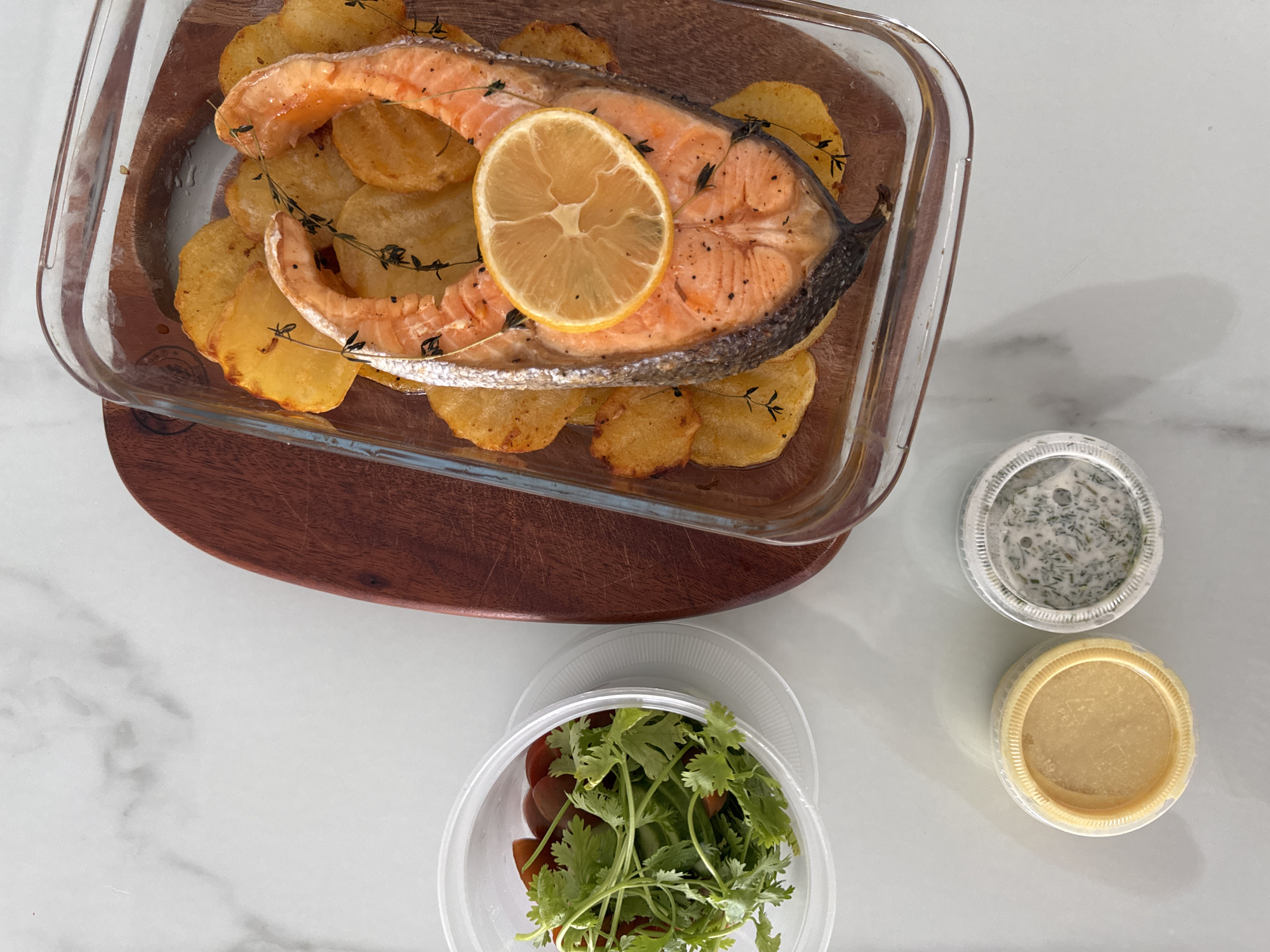 The image depicts a prepared meal on a white marble surface. A rectangular glass dish containing a salmon fillet atop sliced potatoes rests on a wooden board. A slice of lemon and sprigs of thyme adorn the salmon. To the right of the dish are two small, round containers of sauce. Below the glass dish, there is a plastic container of fresh herbs and sliced tomatoes. Based on the provided location information, the image was taken in Bac Nha Trang Ward, Vietnam.