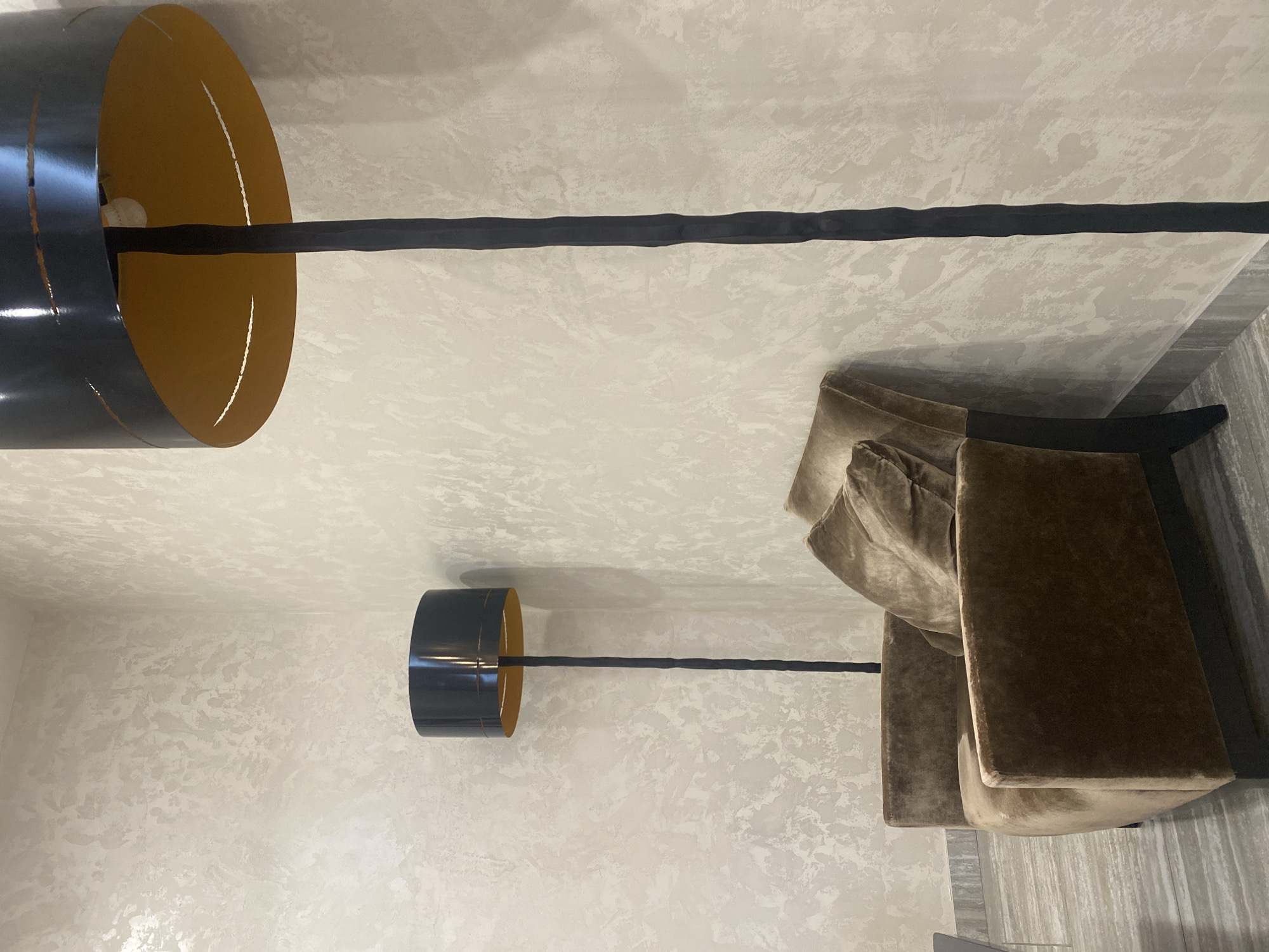 The image displays an interior scene within a room. The walls have a textured, off-white finish. In the foreground, a tall, black floor lamp with a black cylindrical shade and a gold interior is prominently featured, positioned on the right side of the frame. To the left of this lamp, a smaller, similar floor lamp is visible. Between the two lamps, a plush, brown armchair with a matching throw pillow is placed. The armchair is upholstered in a velvet-like fabric. The flooring appears to be a light-colored material with a striped pattern, possibly wood or tile. The scene suggests a modern, minimalist interior design aesthetic. The image does not contain any identifiable people or explicit actions, but rather depicts a static arrangement of furniture and lighting within a domestic or commercial space. No specific details regarding the location in Minsk, Belarus are provided within the visual information of the image.