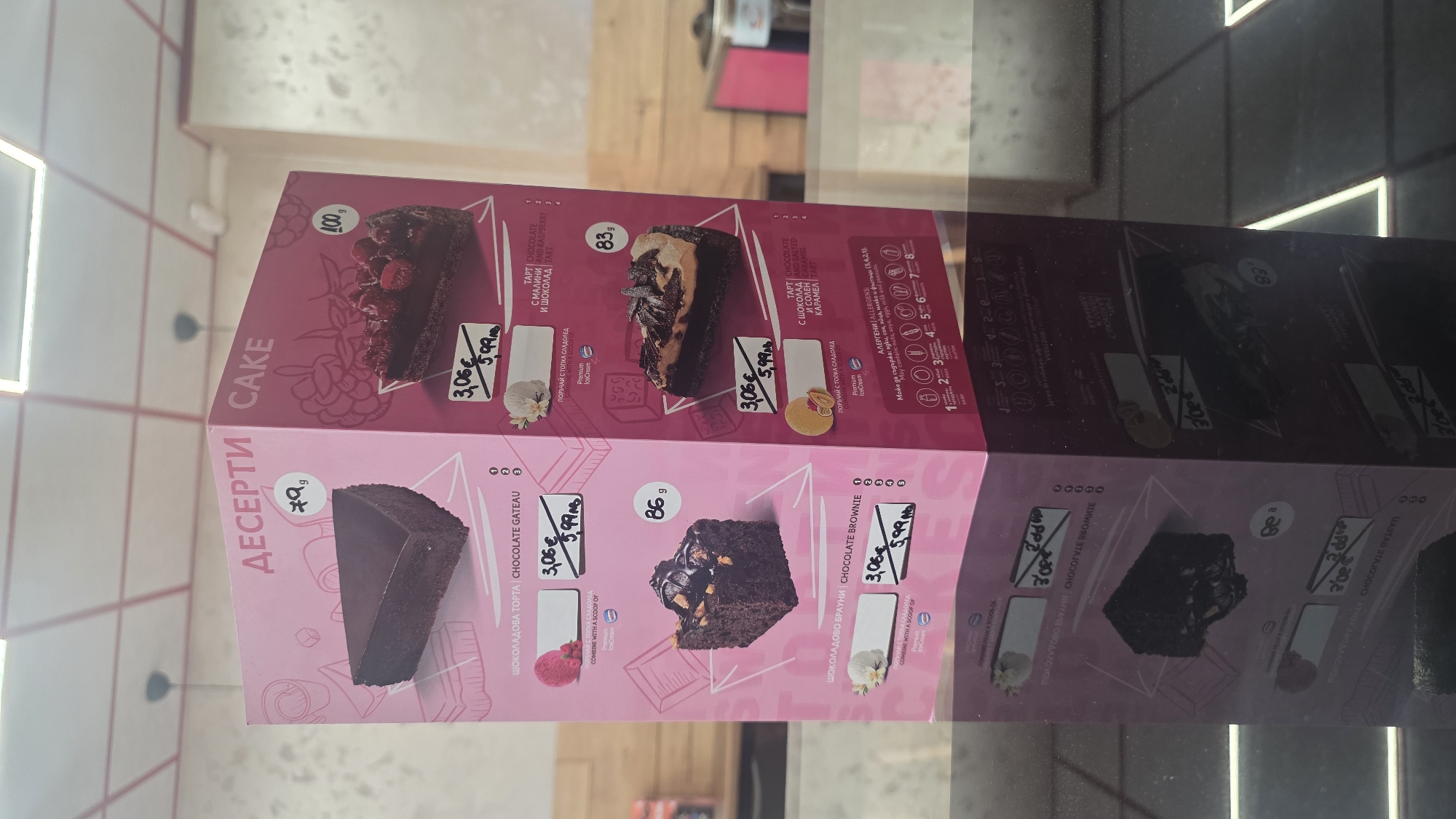 The image shows a cake menu, presumably displayed in a shop or cafe selling desserts. The menu lists several types of cakes, including chocolate gateau, chocolate brownie, chocolate and raspberry tart, and chocolate with salted caramel tart. Each item has a picture, the weight (in grams), and two prices - 3.06€ and 5.99€. The menu is written in Bulgarian and English. It also mentions the possible presence of allergens such as nuts, soya, eggs, milk, and peanuts. The background is blurred, suggesting the menu is displayed indoors, perhaps in a shop window. There's no evidence to pinpoint the exact location in Burgas, Bulgaria.