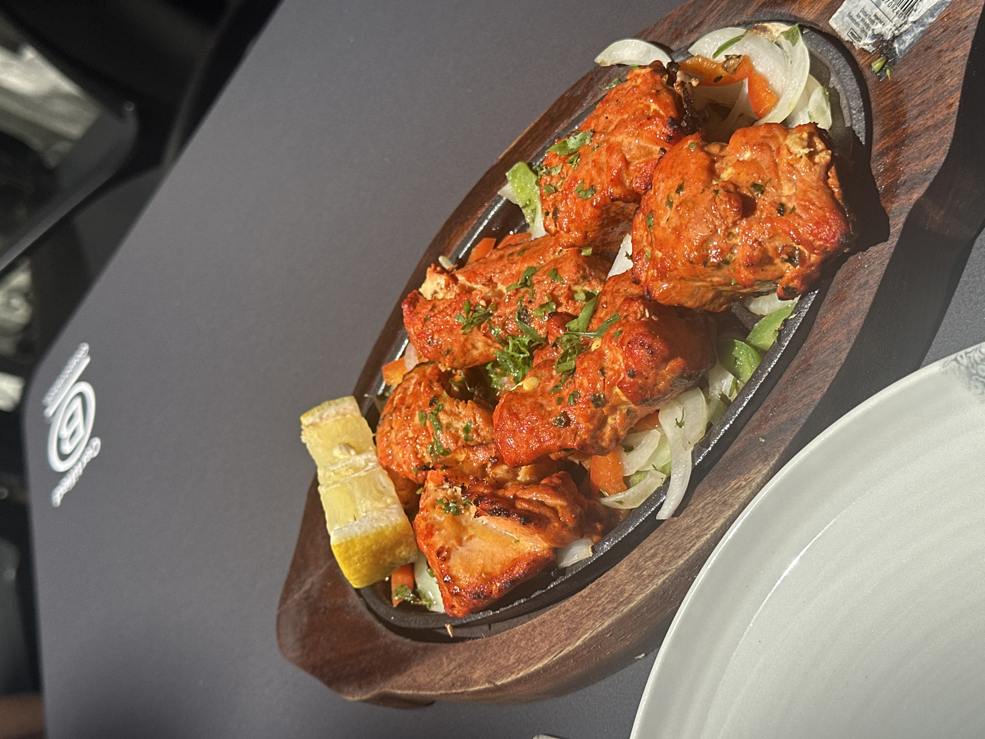 A top-down, angled shot presents a plate of Indian cuisine, likely chicken tikka, served on a sizzling platter atop a wooden base. The bright red chicken pieces are garnished with chopped greens and accompanied by lemon wedges and sliced onions. The dish is placed on a dark gray surface, partially obscured by a white plate on the right. A logo with the letter "B" is visible on the dark surface. The location of the image is unclear, but the mention of "Gijón/xixón, Spain" suggests it might be related to a restaurant or dining establishment in that city. There are no people visible in the image.
