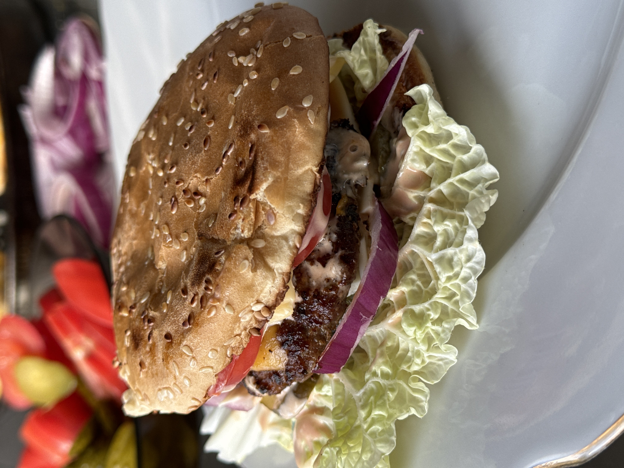 A close-up, eye-level shot focuses on a single hamburger on a white plate. The hamburger is on a bed of crinkled green lettuce. The sesame seed bun is golden brown, and the patty appears thick and well-cooked. Slices of red onion, tomato, and melted cheese are visible beneath the top bun. The hamburger is artfully arranged, with a drizzle of pinkish sauce over the lettuce and some of the ingredients. In the blurred background, hints of red tomato slices, green pickles, and purple onion rings suggest other components of a meal. The lighting is soft, highlighting the textures and colors of the food. There is no indication of the location being in Поназырево, Russia.