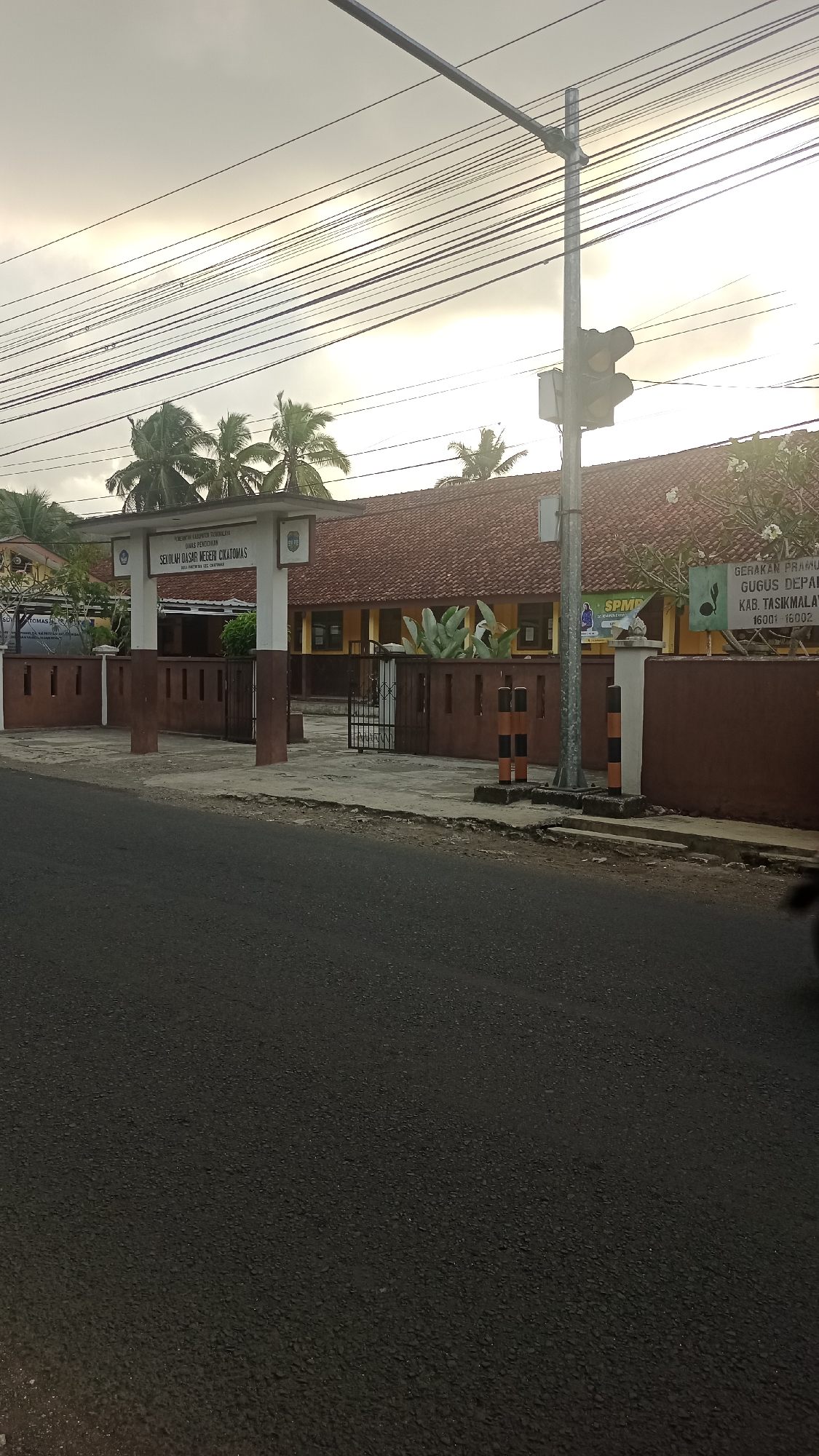This image captures the entrance to SDN Cikatomas, a public elementary school located in Karyamandala, Indonesia. The school building, painted yellow with a red tiled roof, is visible behind a brown-colored wall and gate. A white archway above the gate displays the school's name. To the right of the entrance, a traffic light stands on a pole, and a sign indicates "GERAKAN PRAMUKA GUGUS DEPAN KAB. TASIKMALAYA 16001 16002". Palm trees are visible on the left and right sides of the school. The foreground shows a dark asphalt road, with power lines stretching across the overcast sky. No people are clearly visible in the image, but a portion of a vehicle's side is visible on the far right.