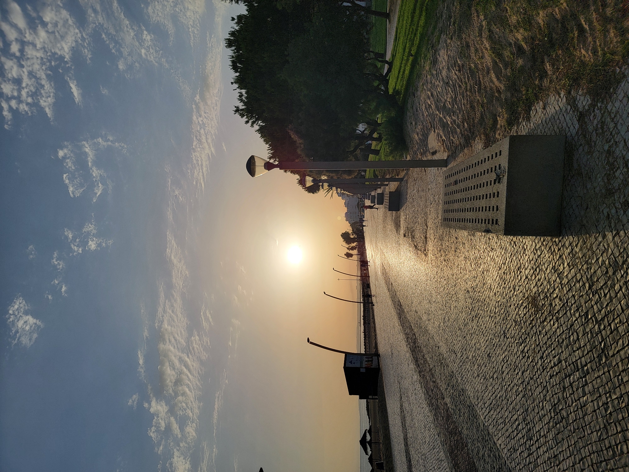 This is a wide-angle, eye-level shot of the promenade in Quarteira, Portugal, at sunset. The sun, a bright orb, is low in the sky, casting a warm, golden light across the scene and creating long shadows. The promenade itself is paved with intricate, light-colored cobblestones. To the left of the promenade, a sandy beach area is visible, with a few thatched umbrellas and a small, dark, cuboid structure. Further down the promenade, several modern streetlights with curved poles and distinctive lantern tops line the walkway. A few scattered individuals are walking or standing along the promenade. To the right of the promenade, a strip of sand leads to a lush green lawn, bordered by a row of dark green, leafy trees. The sky above is a soft blue, with scattered, wispy white clouds that are catching the last rays of the sun.