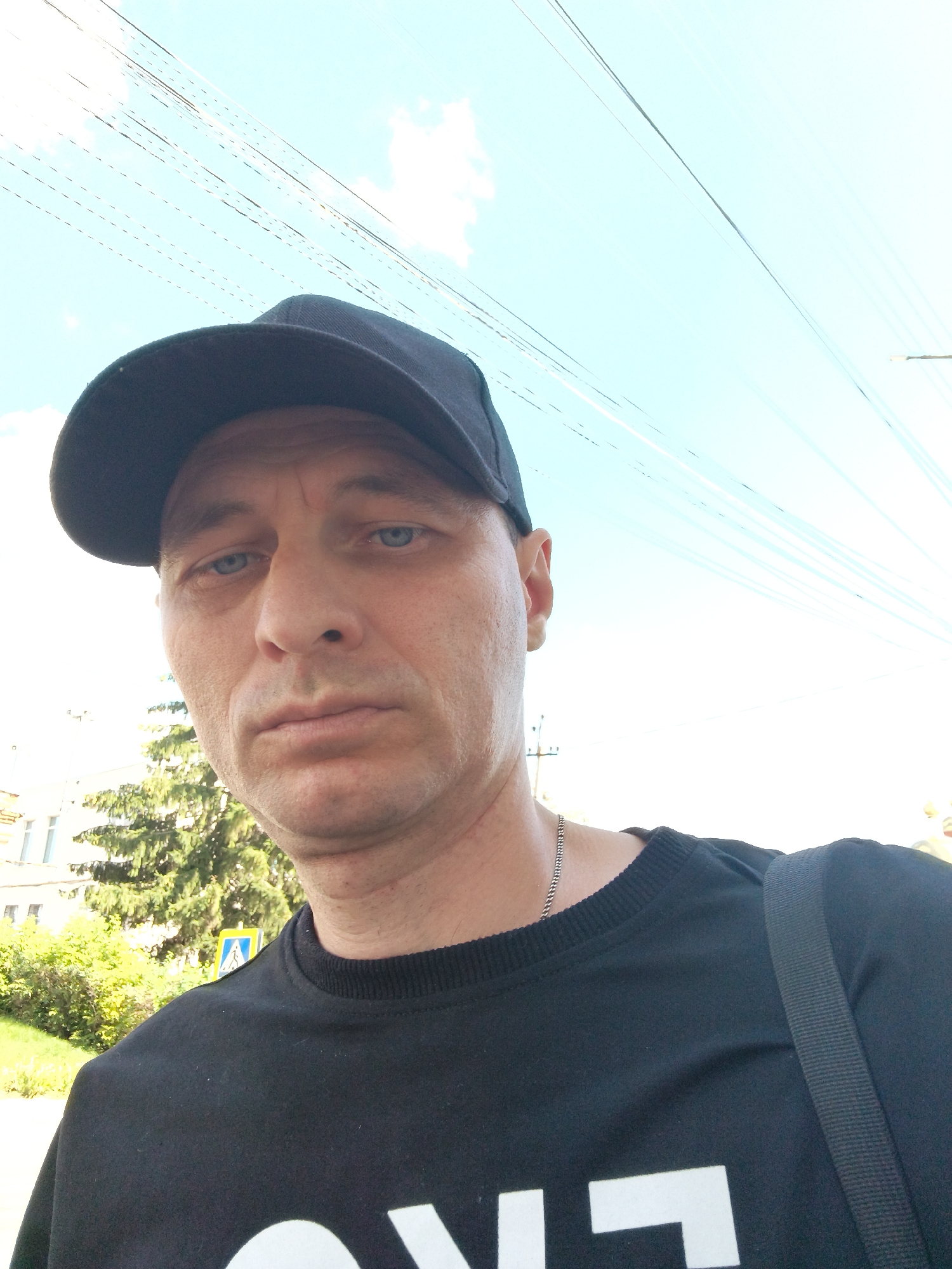 The image shows a selfie of a man wearing a black baseball cap and a black sweatshirt. He has blue eyes and appears to be in his 40s or 50s. He is outdoors, with a clear blue sky and some clouds visible in the background. There are also power lines and trees, along with a building, suggesting an urban or suburban environment. A yellow traffic sign with a blue pedestrian symbol is partially visible behind him, indicating he is likely near a pedestrian crossing. The man is looking directly at the camera with a neutral expression. The photograph seems to have been taken during the daytime.