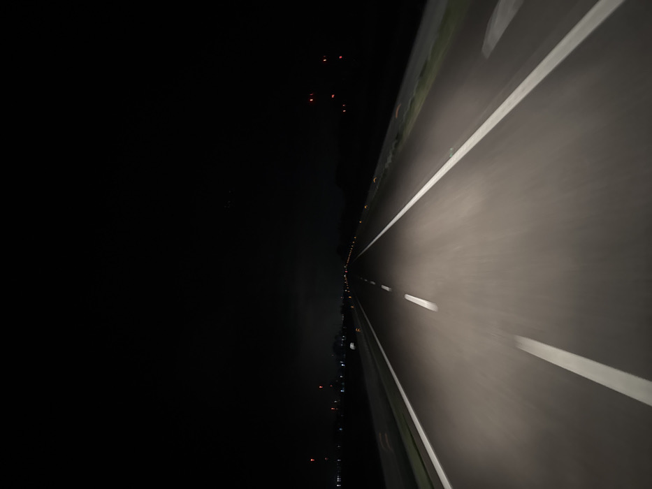 The image displays a multi-lane highway at night, observed from the driver's perspective of a moving vehicle. The scene is located on a road within or directly adjacent to Foggia, Italy. The road surface is illuminated by headlights, revealing an asphalt pavement with prominent white markings: a dashed white line indicating a lane separator in the center-left, and a solid white line defining the right shoulder. Numerous yellow-orange reflective road studs are visible along the right shoulder line, receding into the distance.

The sky overhead is dark, consistent with nighttime conditions. To the left of the highway, beyond the immediate roadside, a dark expanse is punctuated by scattered distant lights, including several red lights and other faint illuminations, suggesting an urban or industrial periphery. Multiple tall structures topped with red warning lights are discernible in the far distance on the left, alongside one distinct bright white object. To the right of the highway, a dark, undeveloped area is visible beyond a guardrail or barrier, with minimal distant light sources. No individuals, explicit subjects, or direct interactions are observable. The foreground of the road exhibits motion blur, indicative of vehicular movement.