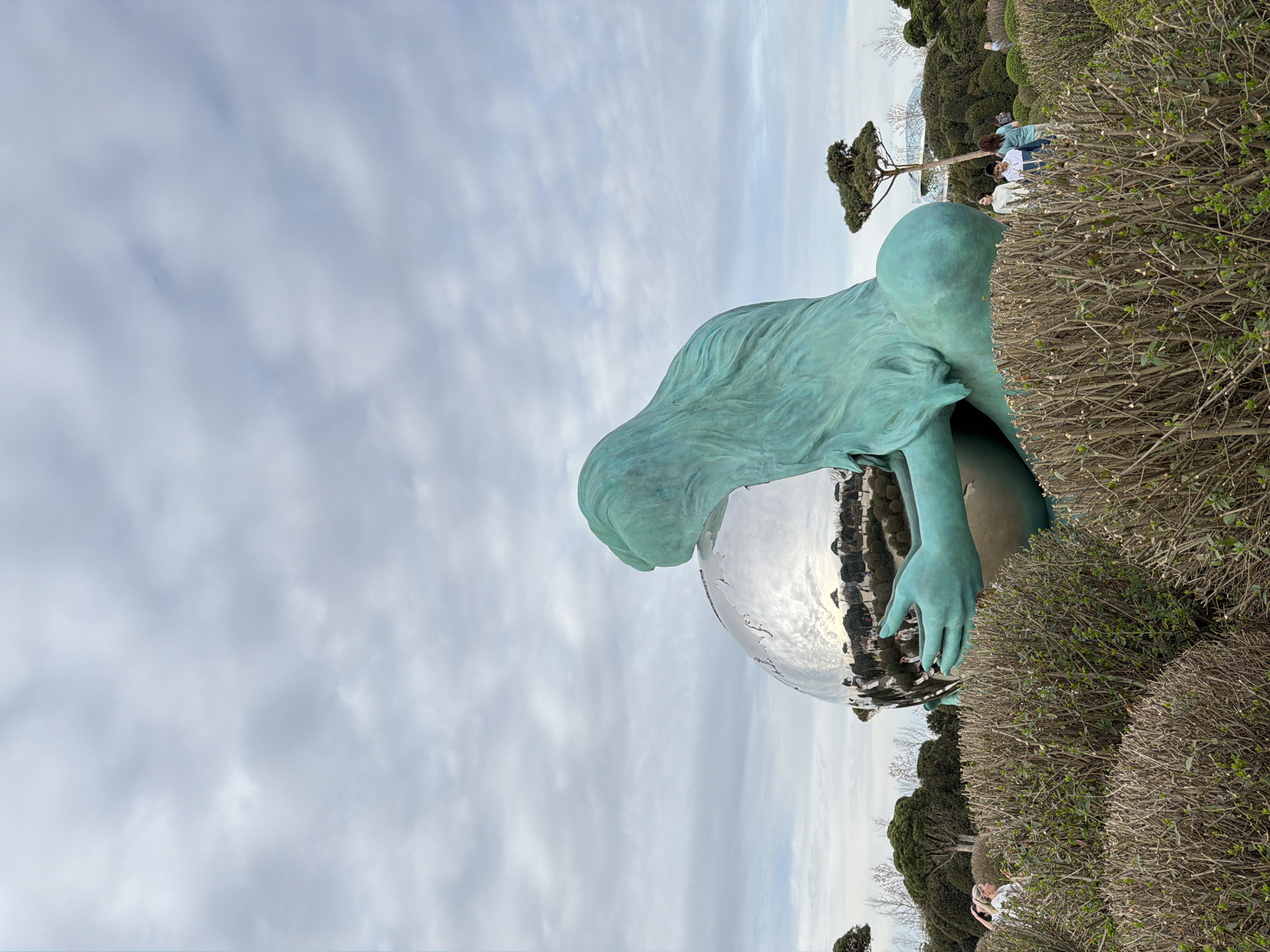 The image shows a statue in Krasnodar, Russia. The statue is of a woman, colored in a blueish-green hue, embracing a large, silver globe. The statue is partially obscured by manicured hedges. In the background, there are people and trees. The sky is overcast with light grey clouds.