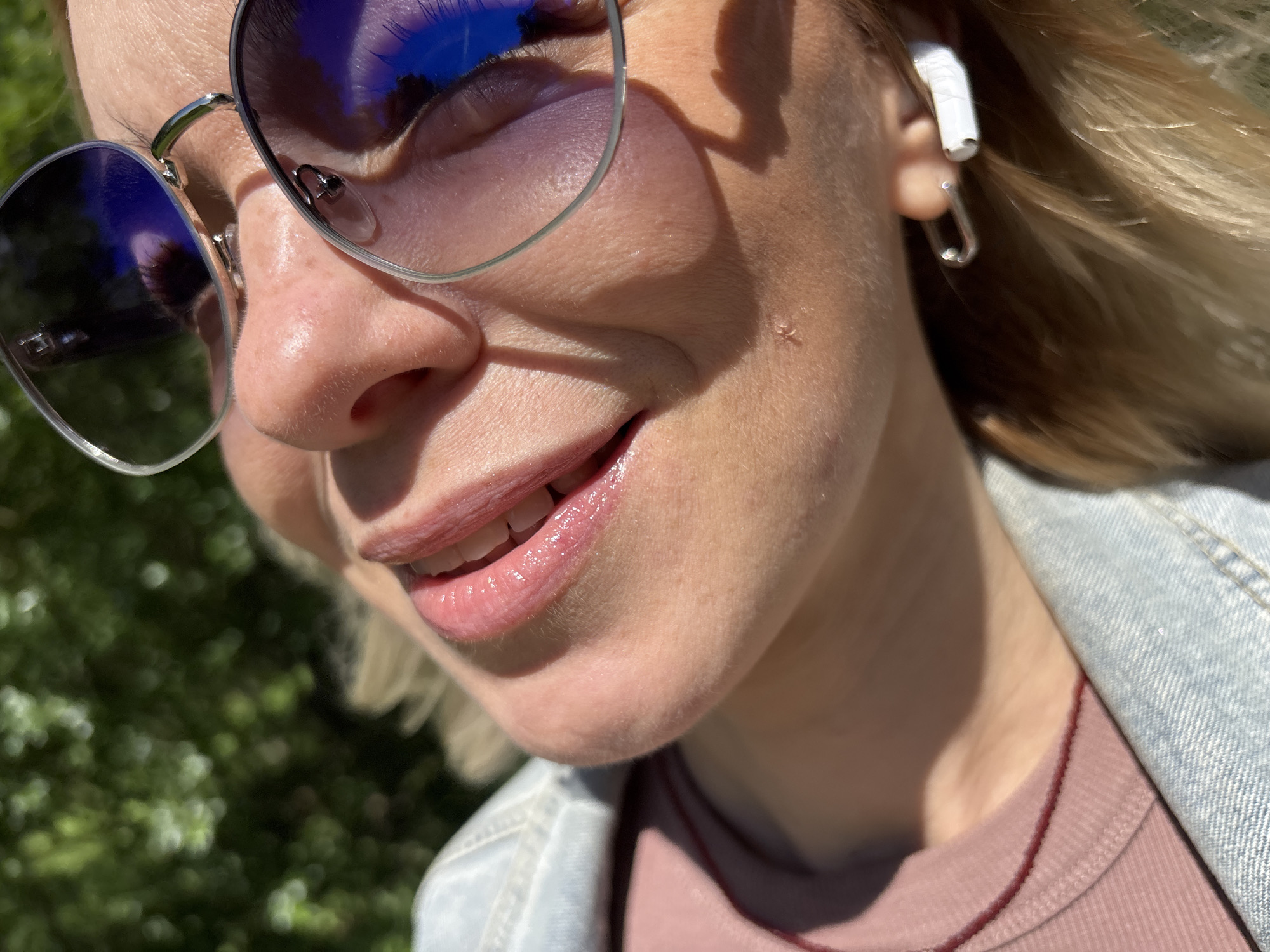 This is a close-up, eye-level, outdoor shot of a person's face, focusing on their mouth and nose, with a portion of their ear and shoulder also visible. The person is wearing large, aviator-style sunglasses with reflective lenses that show a blue sky with some cloud reflection. They are also wearing white wireless earbuds, with one earpiece in their ear and a silver, paperclip-shaped earring hanging from it.
The person has fair skin, with some visible pores and a small mark or mole on their cheek. Their lips are slightly parted, revealing teeth, and appear to be glossed, with a subtle pinkish hue. They are smiling or about to smile, suggesting a lighthearted or happy emotion.
The background is a blur of green foliage, indicating an outdoor setting, likely a park or garden, with dappled sunlight creating shadows on the person's face and hair. The lighting suggests it is daytime, likely late morning or afternoon, with bright sunshine. The person is wearing a light blue denim jacket over a pinkish-toned top, with a dark red string necklace visible.
The image does not contain any visible text. The setting is identified as Saint Petersburg, Russia, but there are no specific visual cues in the image to confirm this location.