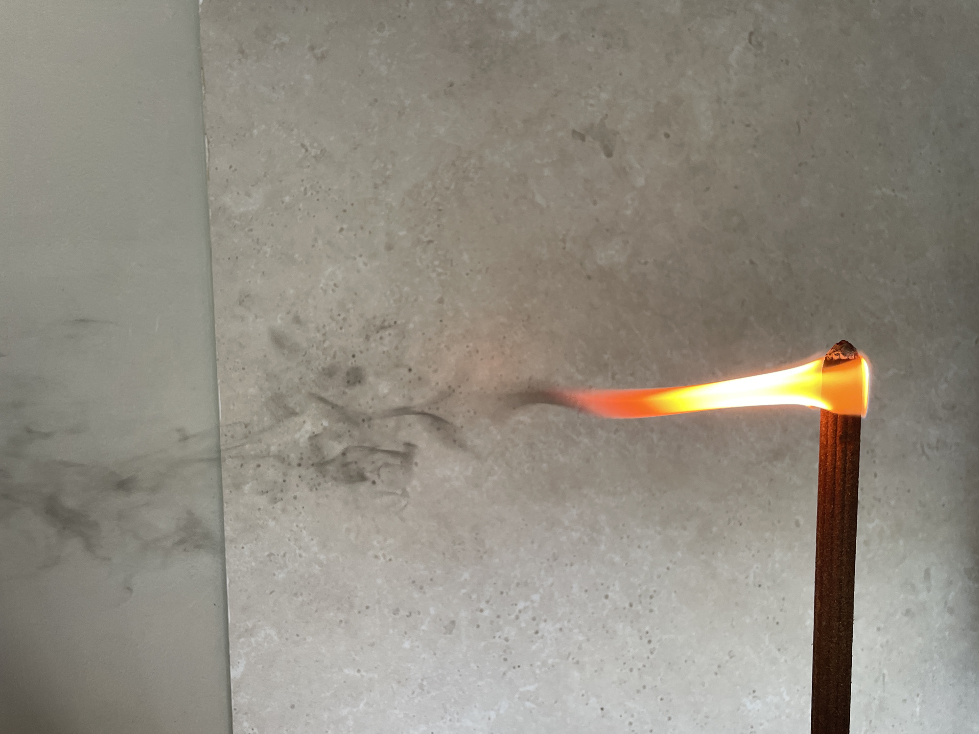 The image shows a close-up, eye-level view of an incense stick that is burning. The stick is dark brown and cylindrical. The tip of the stick is glowing orange and yellow, indicating it is aflame. A plume of grey smoke is rising from the burning tip and dissipating against a light-colored, textured surface, possibly tile or stone. The background is out of focus but appears to be a wall or surface with a marbled or speckled pattern. There is no visible indication of the location within the city of Cahul, Moldova. The scene is static and shows no people or other living beings.