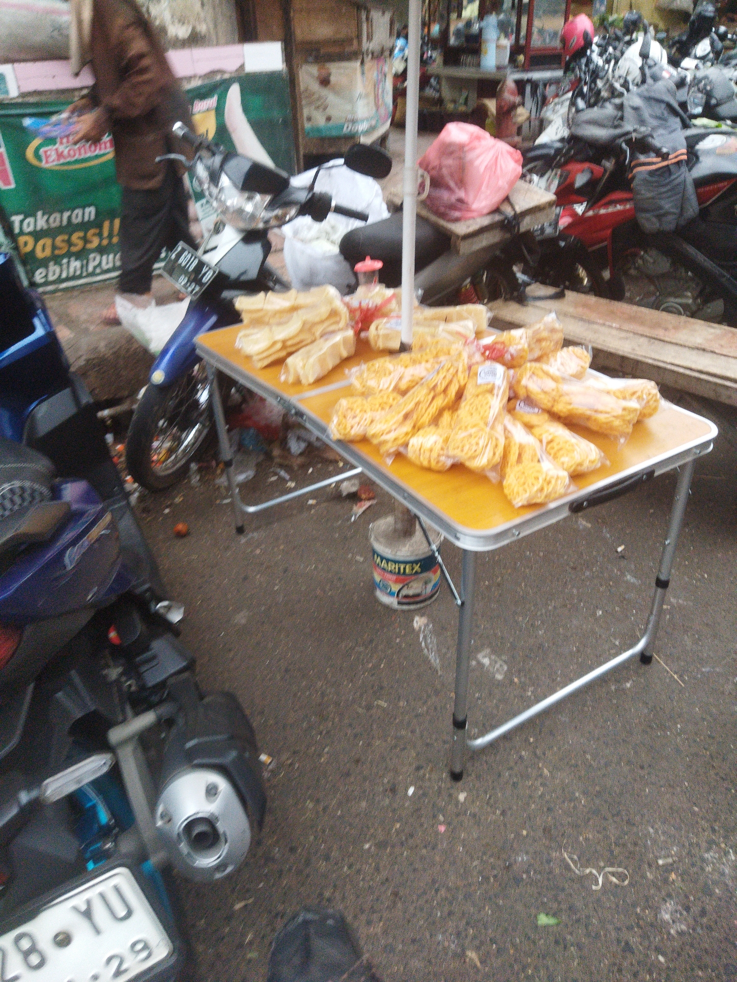 An outdoor, street-level view captures a vendor's stall at what appears to be a local market or roadside establishment in Banjar, Indonesia. In the foreground, a bright yellow folding table is laden with an assortment of packaged snacks, predominantly yellow and appearing to be extruded corn-based snacks or crackers. The snacks are neatly arranged in clear plastic bags. Underneath the table, a cylindrical container labeled "MARITEX" is visible, along with scattered debris and a white plastic bag.
To the left of the table, a blue motorcycle is parked, its front wheel and part of the exhaust system visible. A license plate reading "28-YU 29" is attached to the rear of another motorcycle in the far left, partially obscured. Behind the table, a person dressed in dark clothing, including a head covering and pants, stands with their back to the camera, holding what looks like a stack of snacks or products.
In the background, a green banner with text that includes "Ekonom" and "Takaran Passs!! lebih Pu" is displayed on a wall. Various other motorcycles are parked in the area, some with helmets and other accessories. The overall environment suggests a bustling commercial setting, likely on a street or sidewalk, given the asphalt ground and general clutter. The lighting indicates it is daytime.
