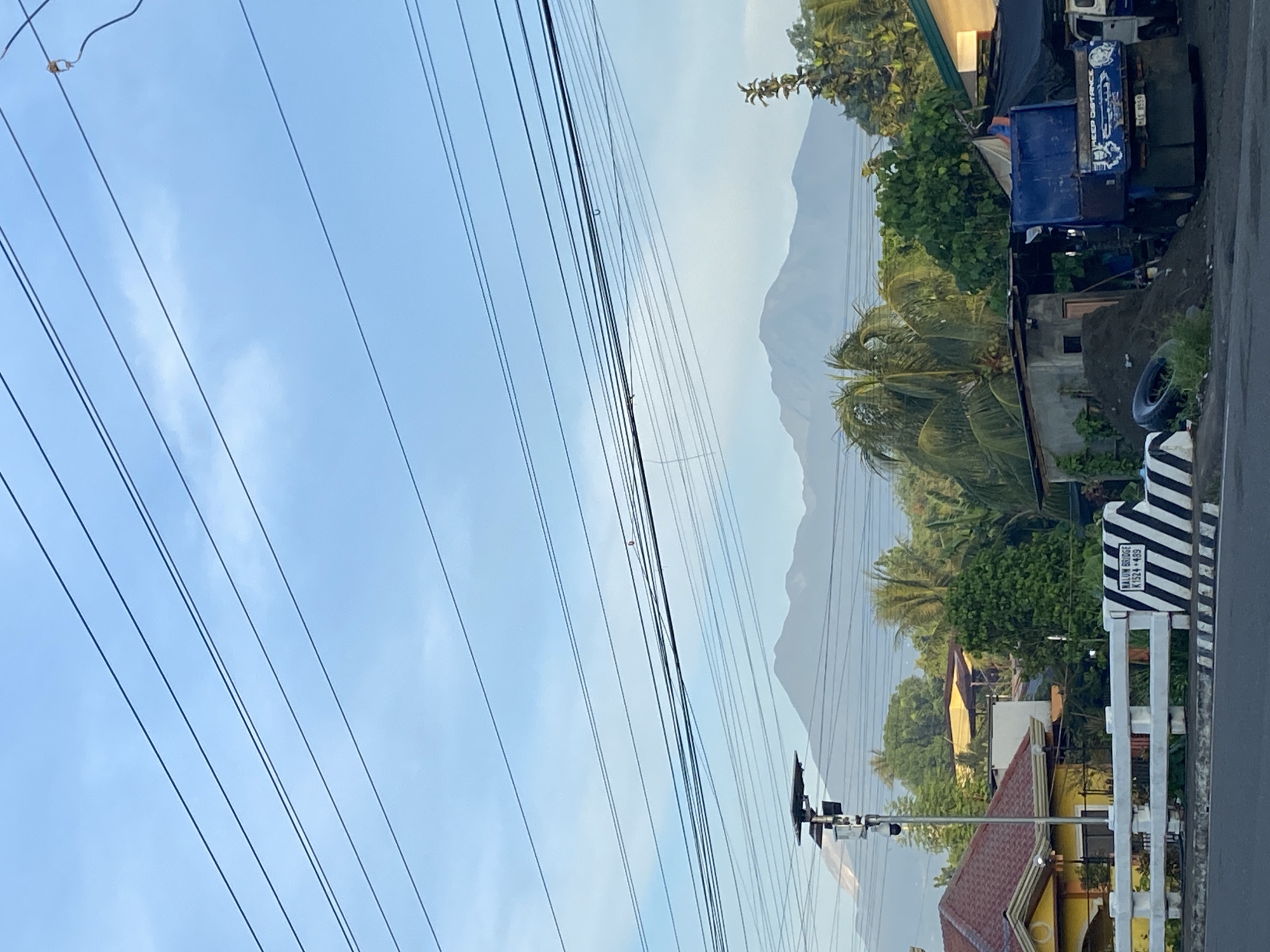 The image depicts a cityscape in Davao City, Philippines, captured on a clear day with light blue sky overhead. Prominent in the scene is a web of overhead power lines that crisscross the sky. A distant, hazy mountain range forms a backdrop, with residential buildings and lush green vegetation in the midground. A blue truck is parked near a building. Street signs and road markings are evident along the roadside. The scene presents a blend of natural landscapes and urban infrastructure.