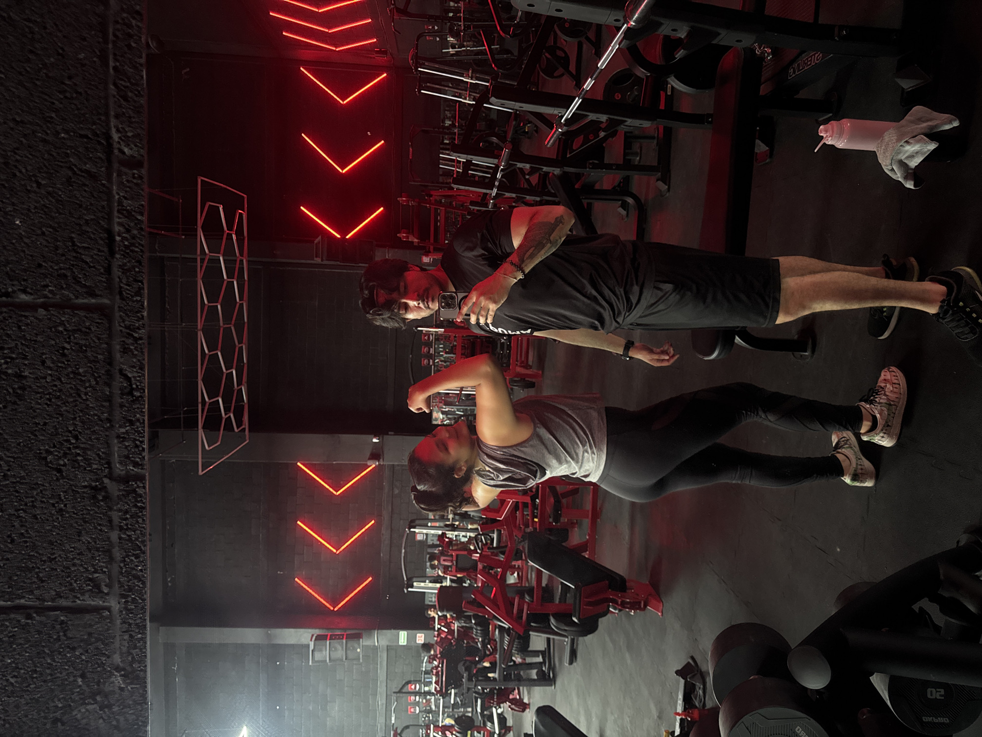Two people are in a gym. The man, wearing a black t-shirt and shorts, is holding a cell phone and appears to be taking a picture. The woman is flexing her bicep. Gym equipment is behind them. Red neon lights are on the wall. The location may be Puebla, Mexico.