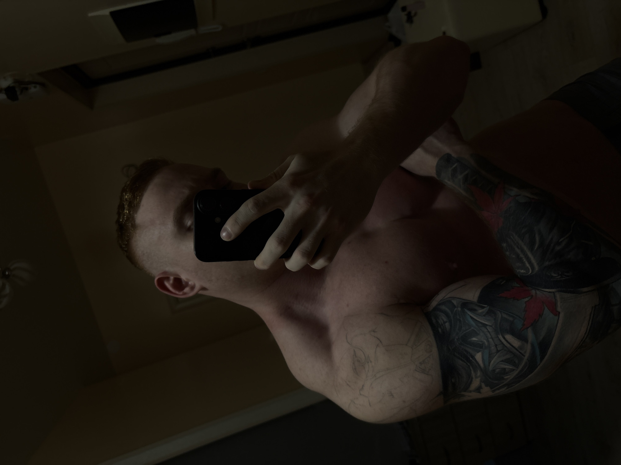 A close-up, dimly lit photograph features a shirtless man with red hair taking a selfie. He is holding a black smartphone in his right hand, obscuring part of his face. The man has tattoos on both arms, with a prominent tattoo of a figure with a red maple leaf on his right arm. The scene appears to be a bathroom, as indicated by a mirror reflecting the room and a light fixture in the background. The environment is dark and slightly grainy. The location of the image is unknown, and there is no clear indication of the city of Chaikovsky, Russia.
