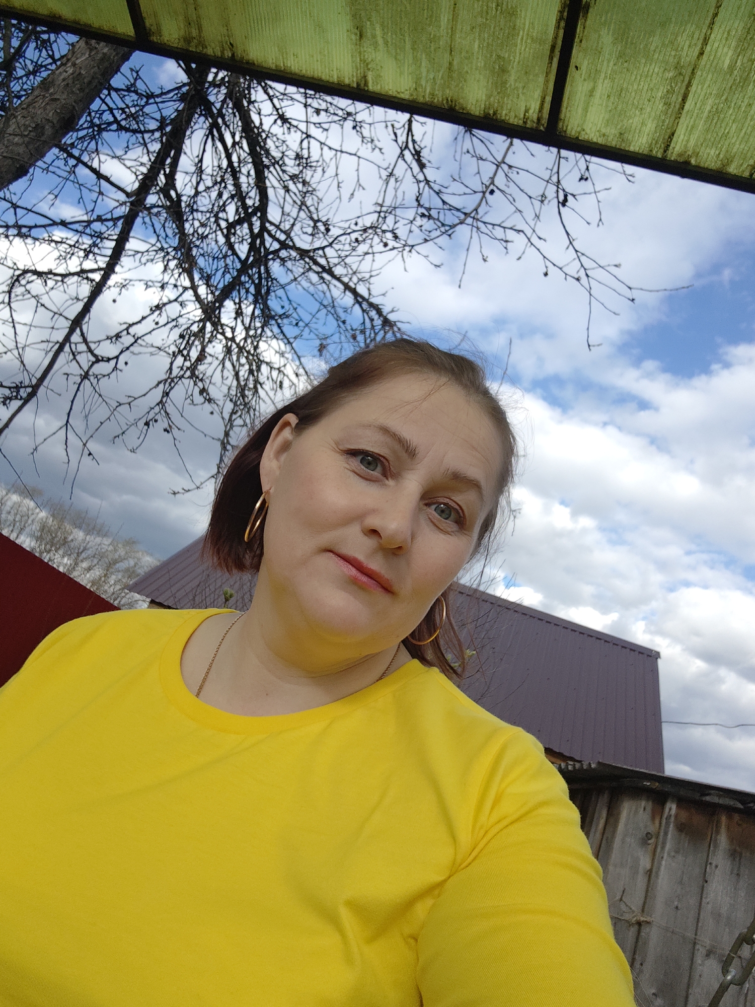 A woman is featured in a close-up, eye-level outdoor portrait, likely a selfie, looking directly at the camera with a soft, pleasant expression and a subtle smile. She has light brown hair, pulled back, and is wearing large gold hoop earrings and a thin gold chain necklace. Her top is a bright, plain yellow long-sleeved t-shirt. She has light makeup, including red lipstick and some eye makeup.
The setting appears to be a backyard or rural area during the daytime, under a partly cloudy sky with visible patches of blue. Bare tree branches crisscross across the upper part of the image, suggesting late autumn, winter, or early spring. At the very top, a section of a green, translucent corrugated plastic roof or awning is visible. In the background, to the woman's left, there's a glimpse of a reddish-brown building or roof. To her right, a larger reddish-brown metal roof structure is prominent, with an older wooden fence visible beneath it. Faint chains can also be seen near the fence on the right. There is no visible text in the image. The overall impression is a casual, relaxed moment captured outdoors in Фоки, Russia.