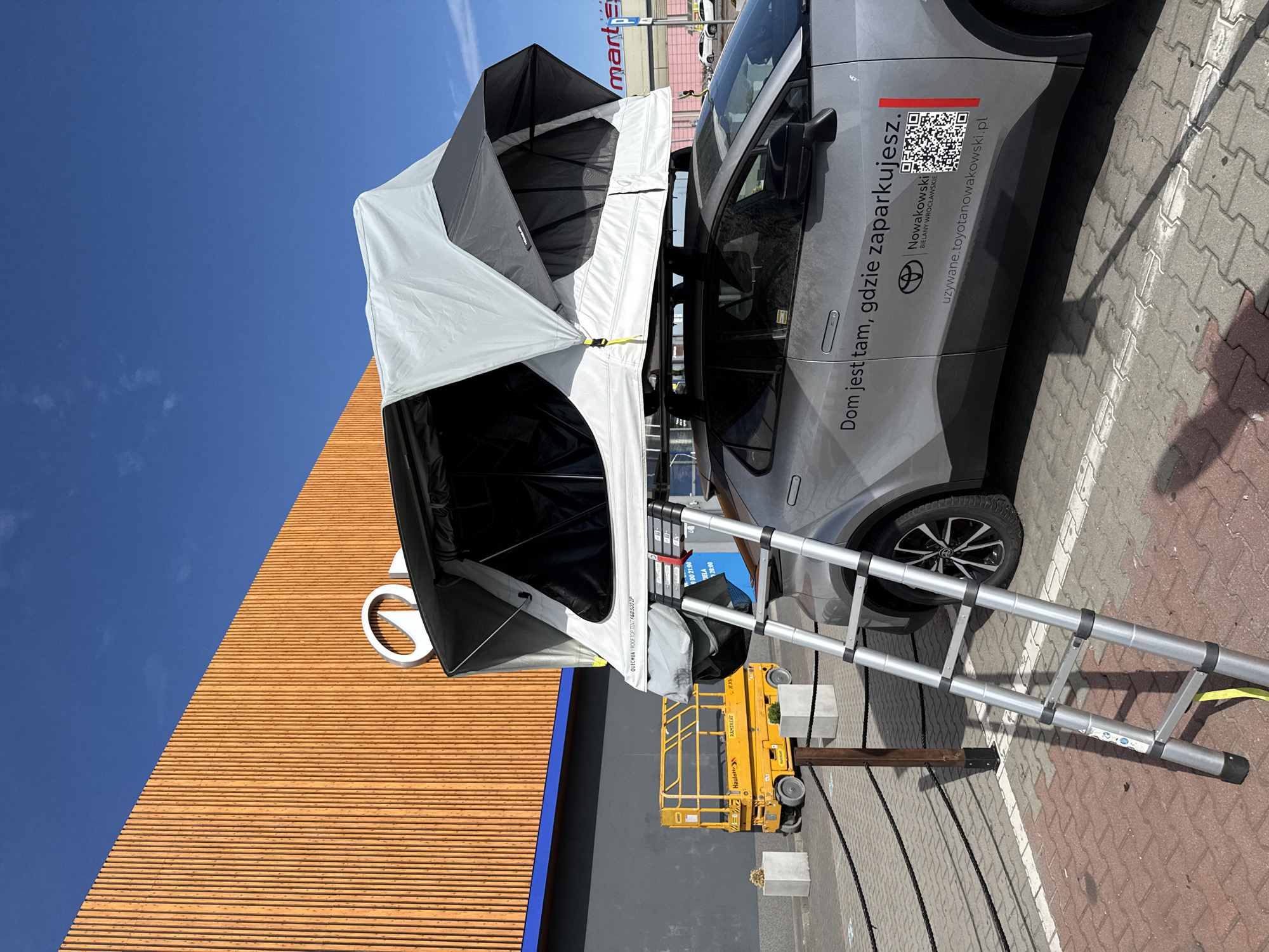 A silver Toyota C-HR is parked in a parking lot outside a building with an orange wooden facade. A rooftop tent is mounted on the car's roof rack, and an aluminum ladder leans against the side of the car, providing access to the tent. The tent is partially open, revealing its interior. In the background, a yellow scissor lift is parked, and a sign with the "mart" logo is visible on the building. The scene appears to be in Wrocław, Poland, as indicated by the text on the car, which reads "Dom jest tam, gdzie zaparkujesz" (Home is where you park) and the website "uzywane.toyotnowakowski.pl," suggesting a Toyota dealership or used car lot. The sky is clear and blue, indicating sunny weather.