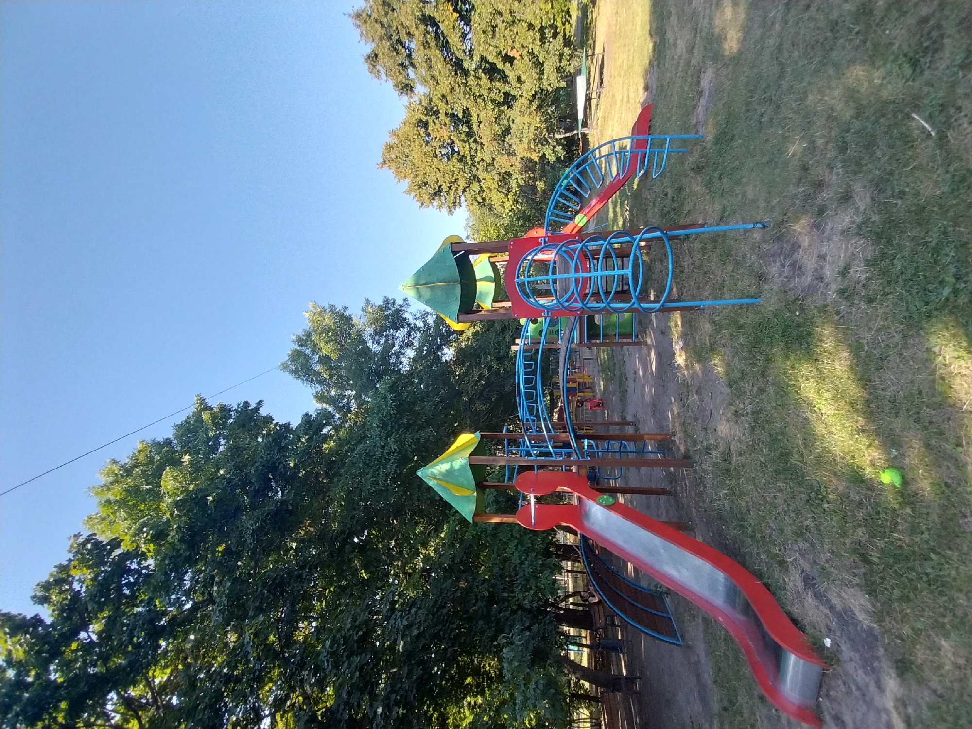 This image depicts a playground in the city of Doslidne, Ukraine. The playground features a multi-colored play structure with red and green slides, blue metal climbing frames, and wooden supports. The area is surrounded by mature trees, casting shadows on the grassy ground. A clear blue sky is visible above. There are benches and a trash can in the background. A single yellow-green ball is visible on the grass in the foreground.