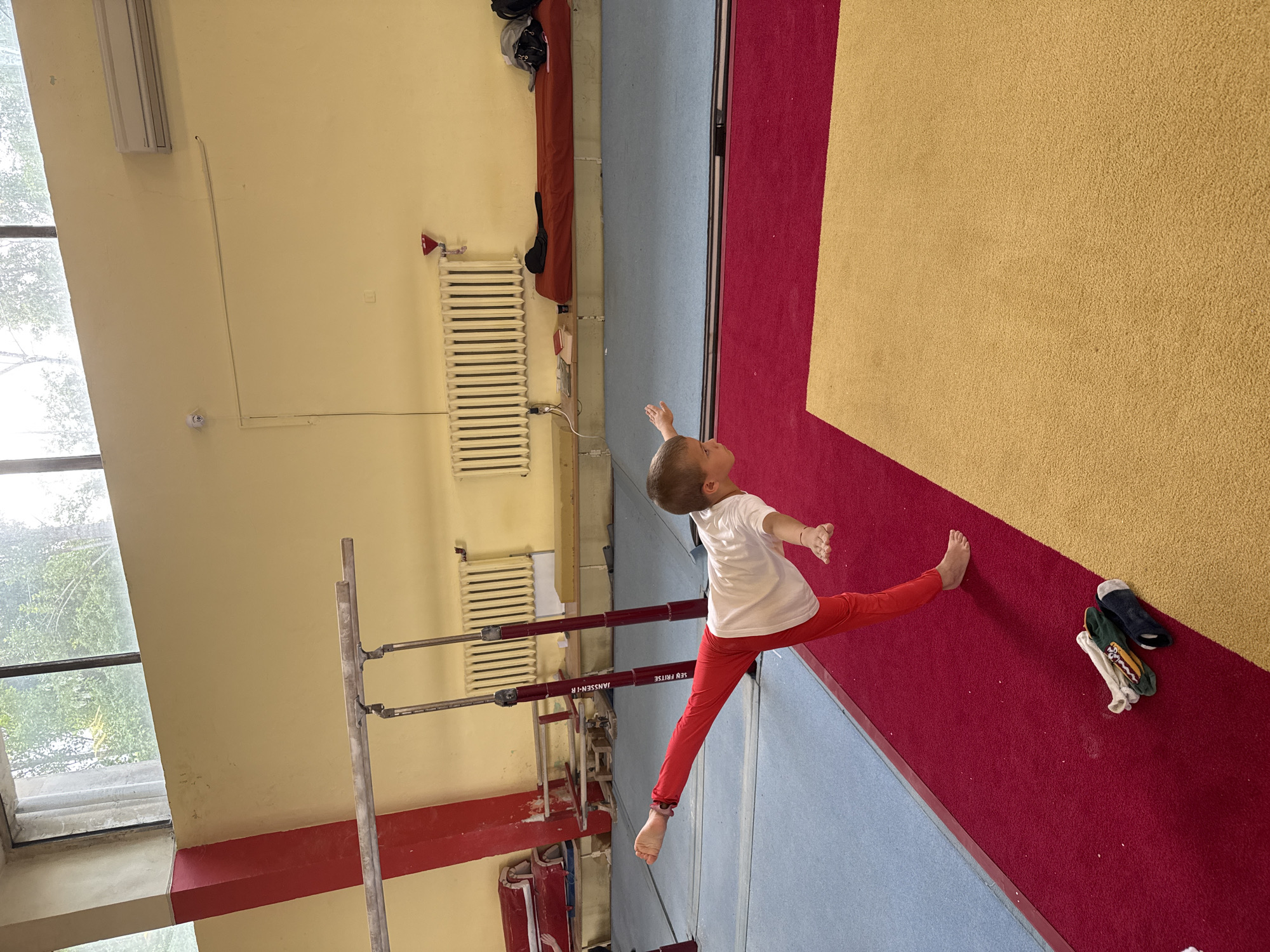 A young child, likely a boy, with short, light brown hair, is captured mid-split in an indoor gymnastics hall. He is wearing a white short-sleeved t-shirt and red pants, with his arms extended outwards and looking upwards and to the right. He is barefoot, demonstrating flexibility on a section of red matting.
The hall features a floor composed of large, distinct sections of red, tan/gold, and blue mats. To the left of the child, a set of uneven bars with metal uprights and red horizontal bars is visible. The brand names "JANSSEN-IK" and "SEB FRITSE" are printed in white text on the red bars. The walls are a light yellow/cream color, with a prominent red horizontal stripe running along the lower section, possibly indicating a protective padding.
On the far left, a large window reveals green foliage outside, suggesting it is daytime. An old-fashioned white radiator is mounted on the wall in the background. Above the radiator, there's a small red triangular sign. A fluorescent light fixture is also visible on the wall near the window. Several pairs of socks are casually placed on the tan/gold mat near the child's right foot. The overall impression is that of a functional, well-used training facility.